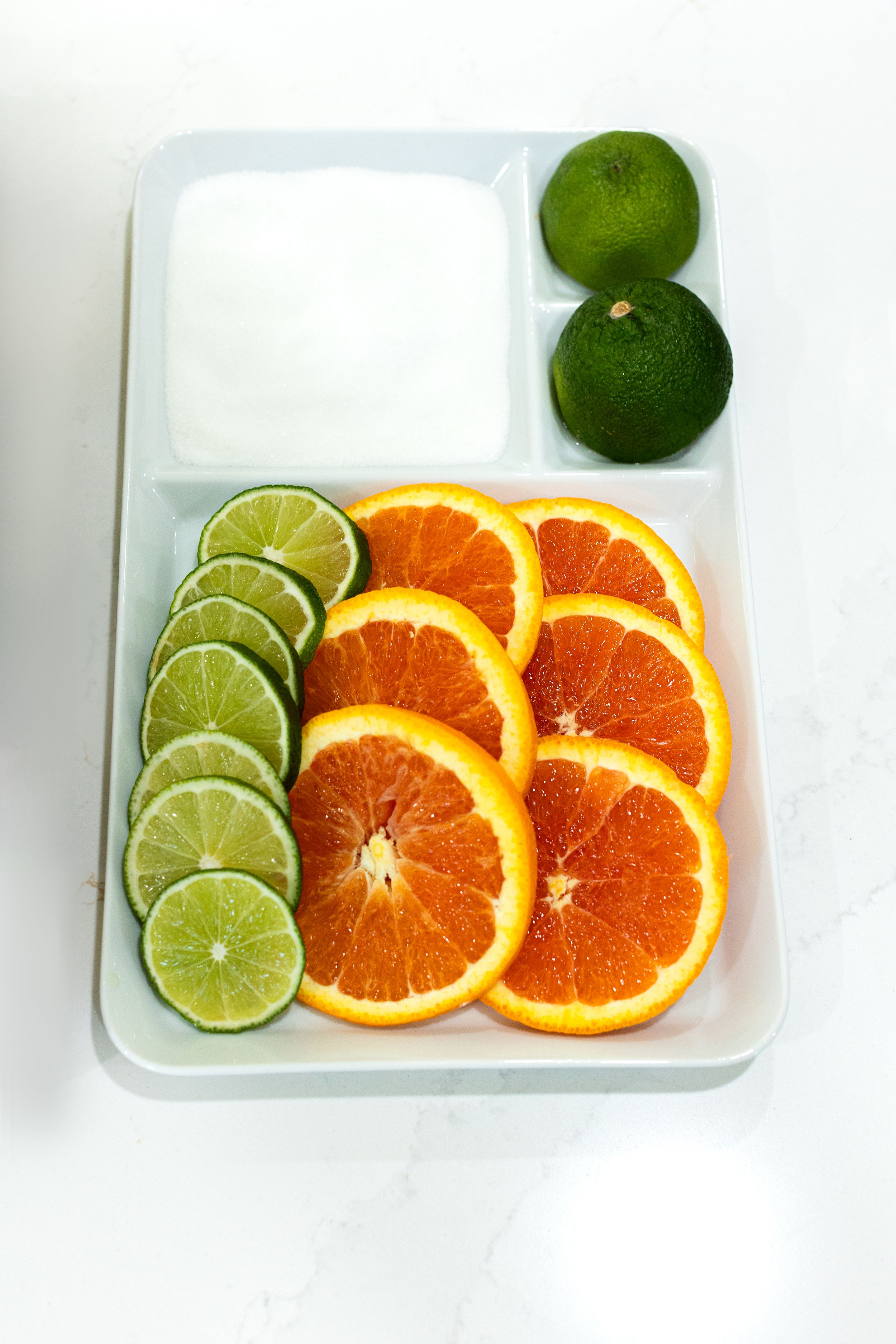 Fresh sliced oranges and limes arranged on a white cutting board with whole limes at the top.