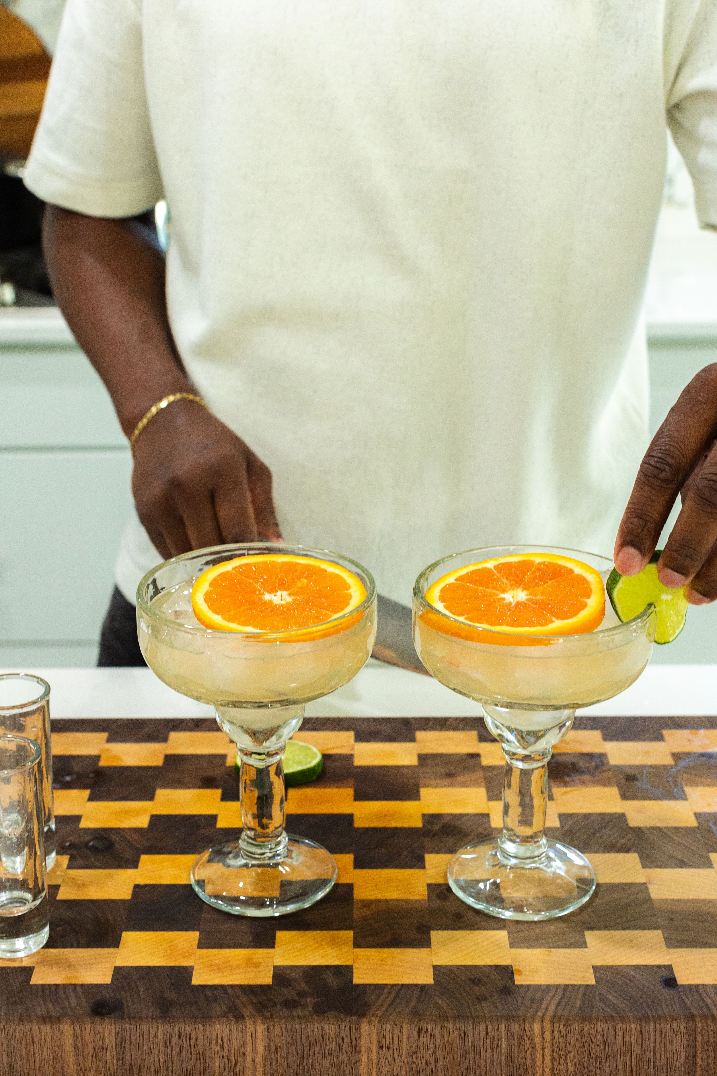 Two orange-garnished cocktails in coupe glasses sit on a wooden checkered cutting board as hands prepare the drinks.