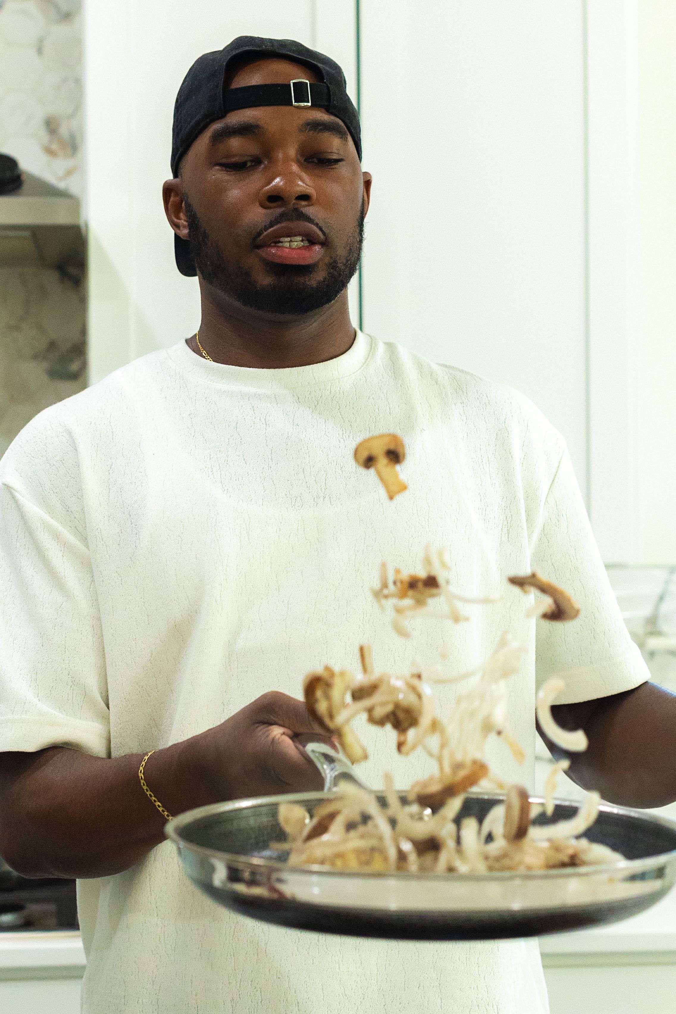 Person in white t-shirt and backwards cap flipping mushrooms in a pan while cooking in a kitchen.