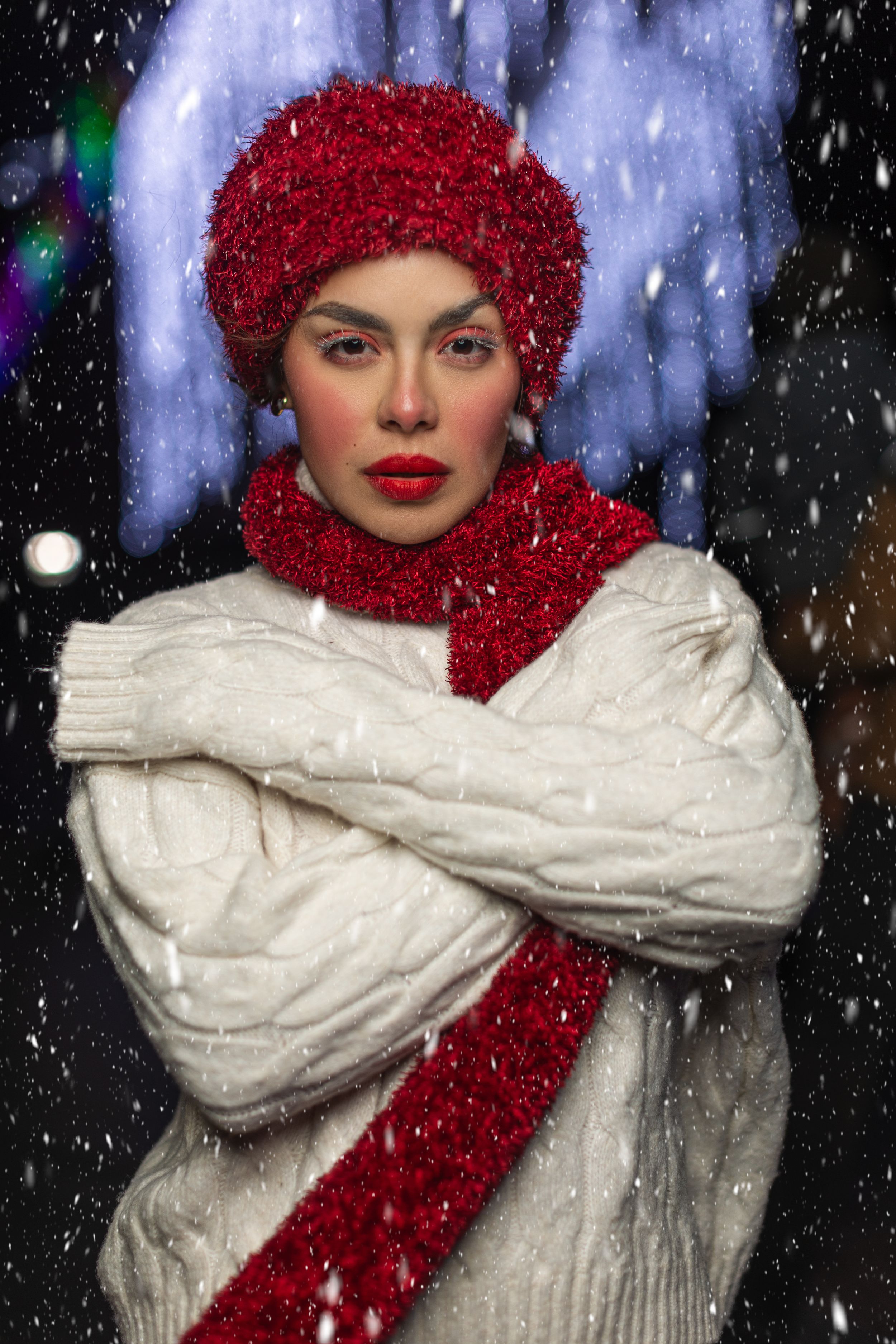 Woman in white knit sweater wearing sparkly red hat and scarf poses in falling snow at night.