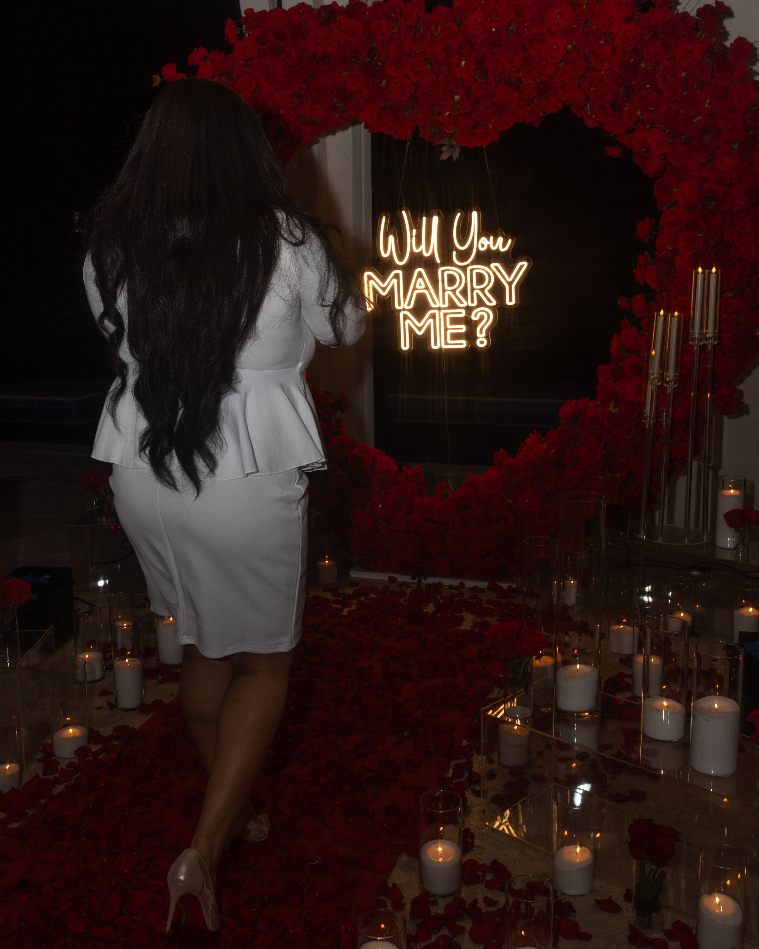 A romantic marriage proposal setup with 'Will You Marry Me' in neon lights surrounded by red roses and candlelight.