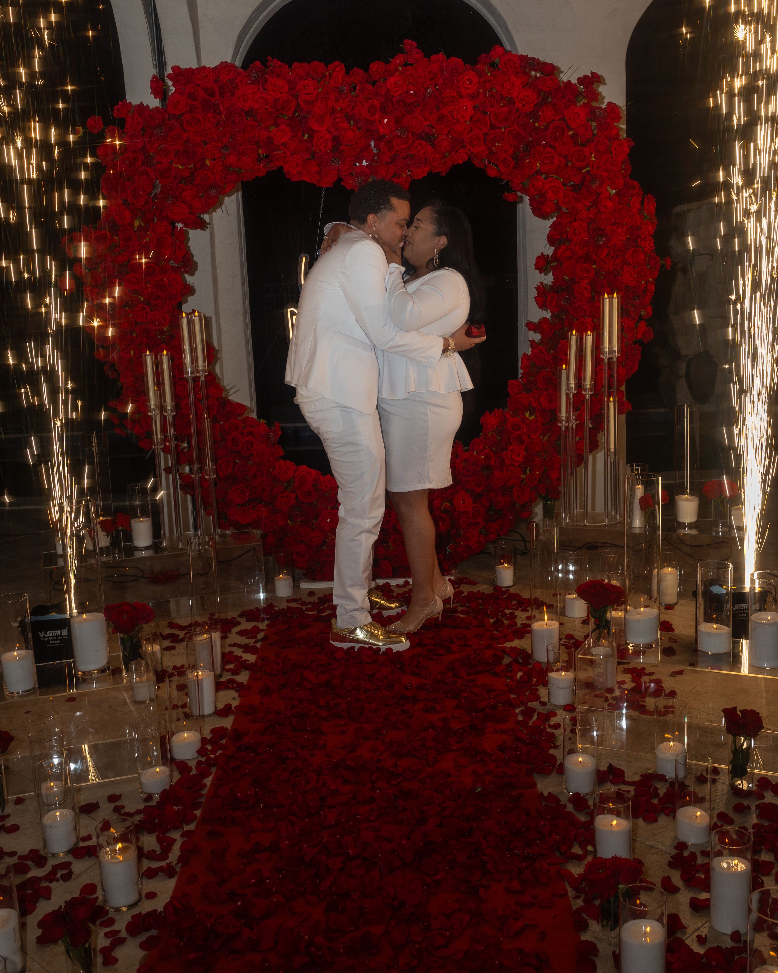 A romantic scene with a pair embracing under a large heart-shaped arch of red roses surrounded by glowing candles on the floor.