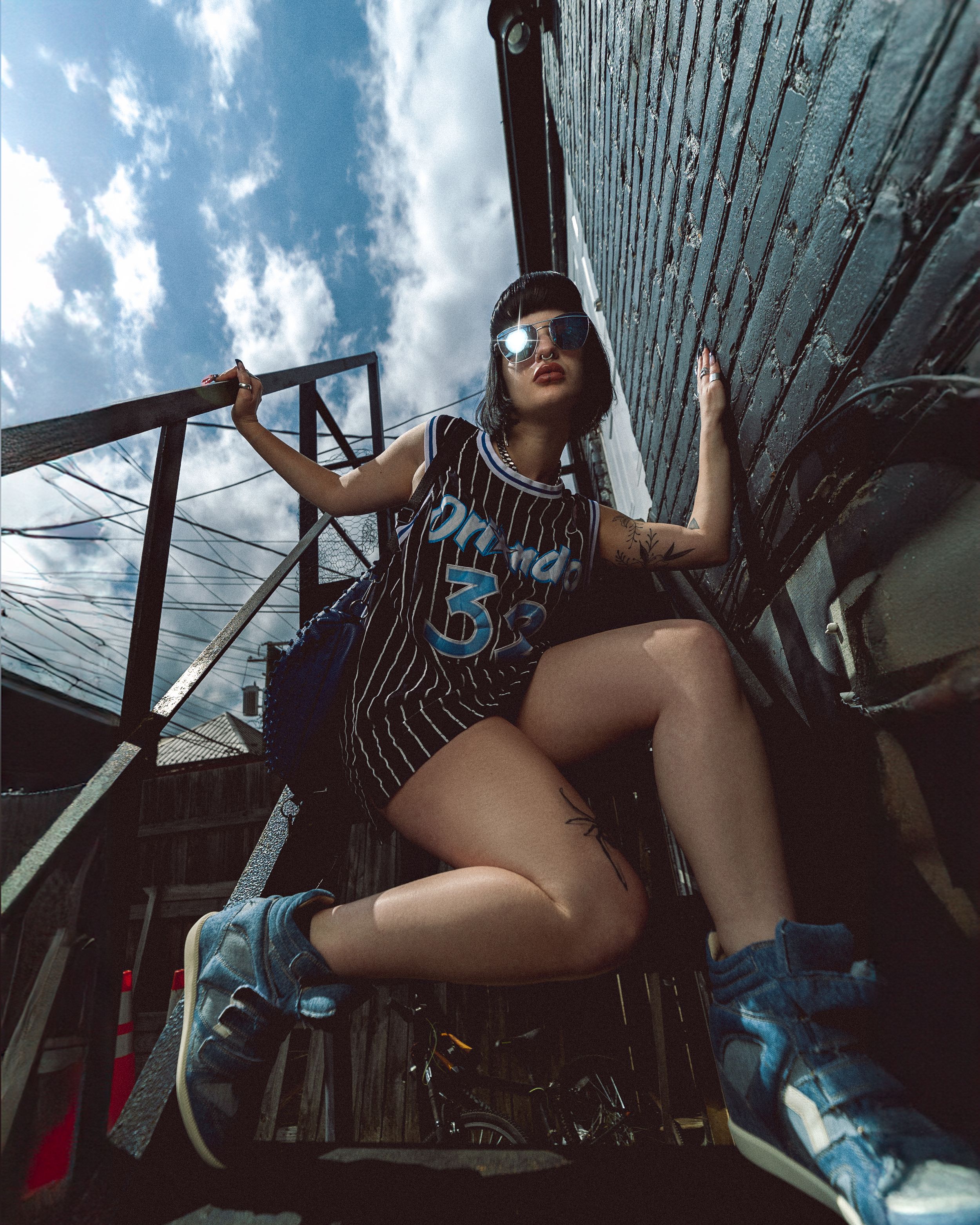 Fashion model in a pinstriped basketball jersey and blue sneakers poses on rustic metal stairs against a cloudy sky.