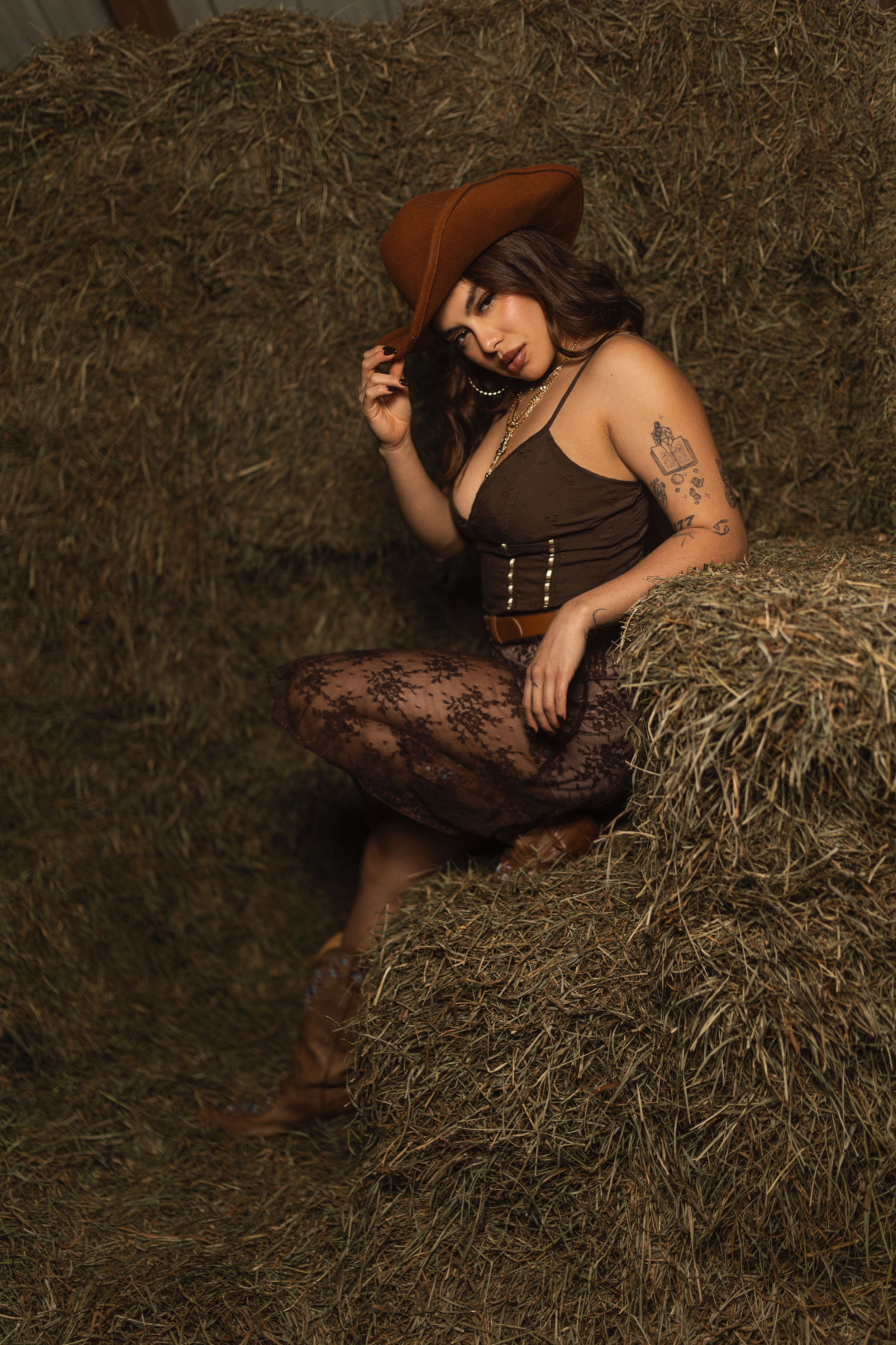 Rustic western-inspired portrait with brown cowboy hat and corset posed against hay bales in moody lighting.