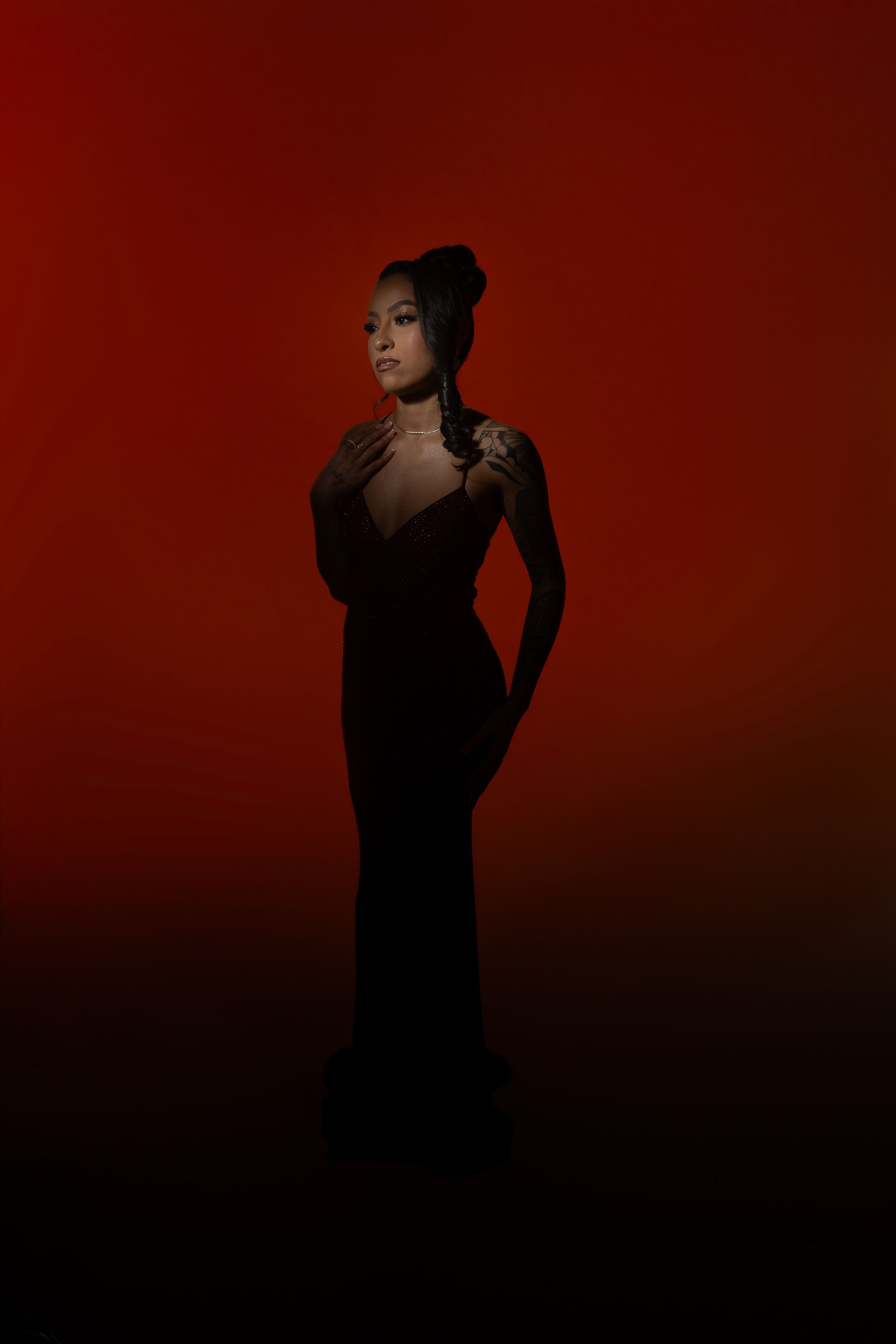 Figure in a black evening gown poses gracefully against a dramatic dark red background in moody lighting.
