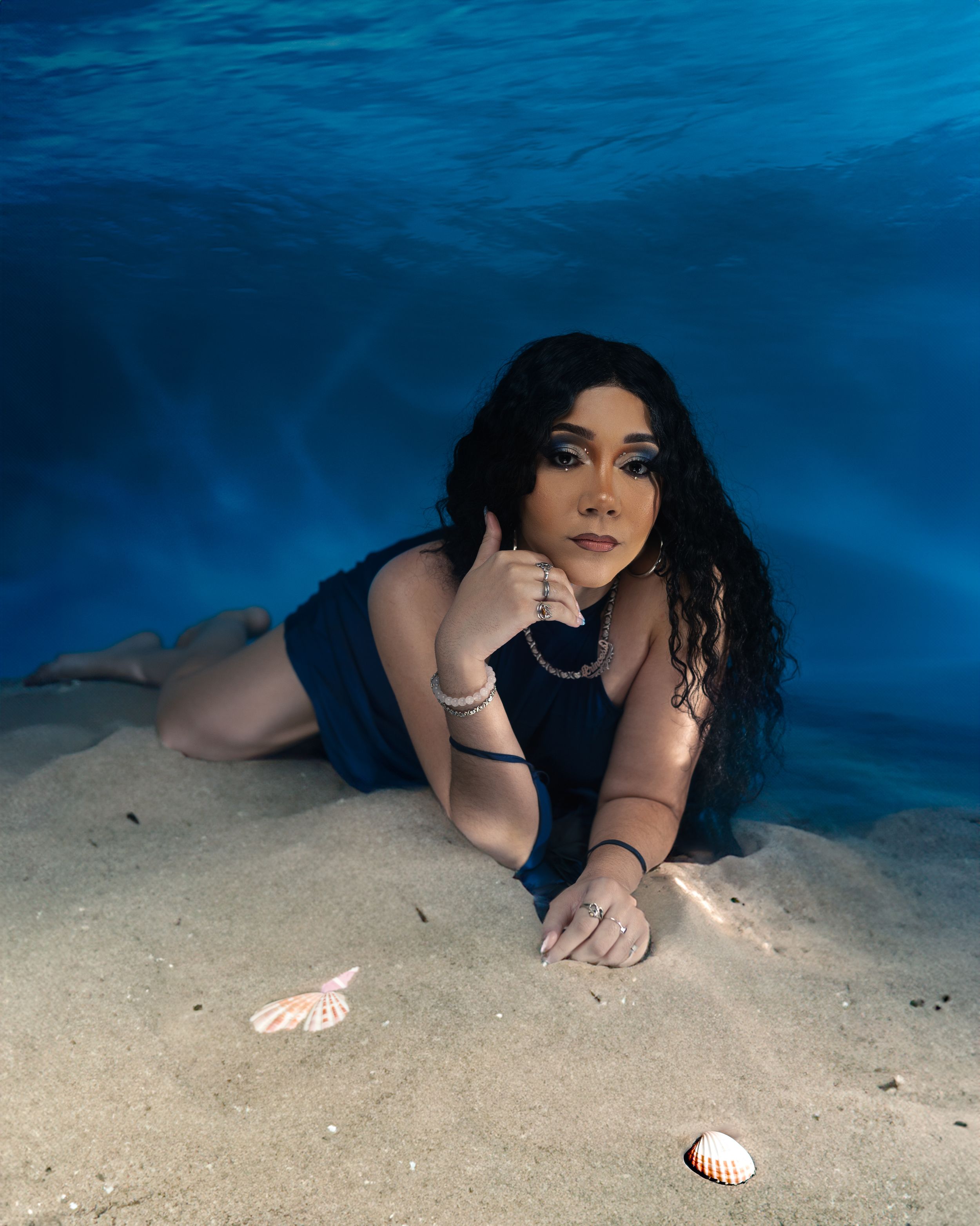 An underwater fashion portrait shows a figure reclining on sandy seabed with seashells nearby in deep blue waters.