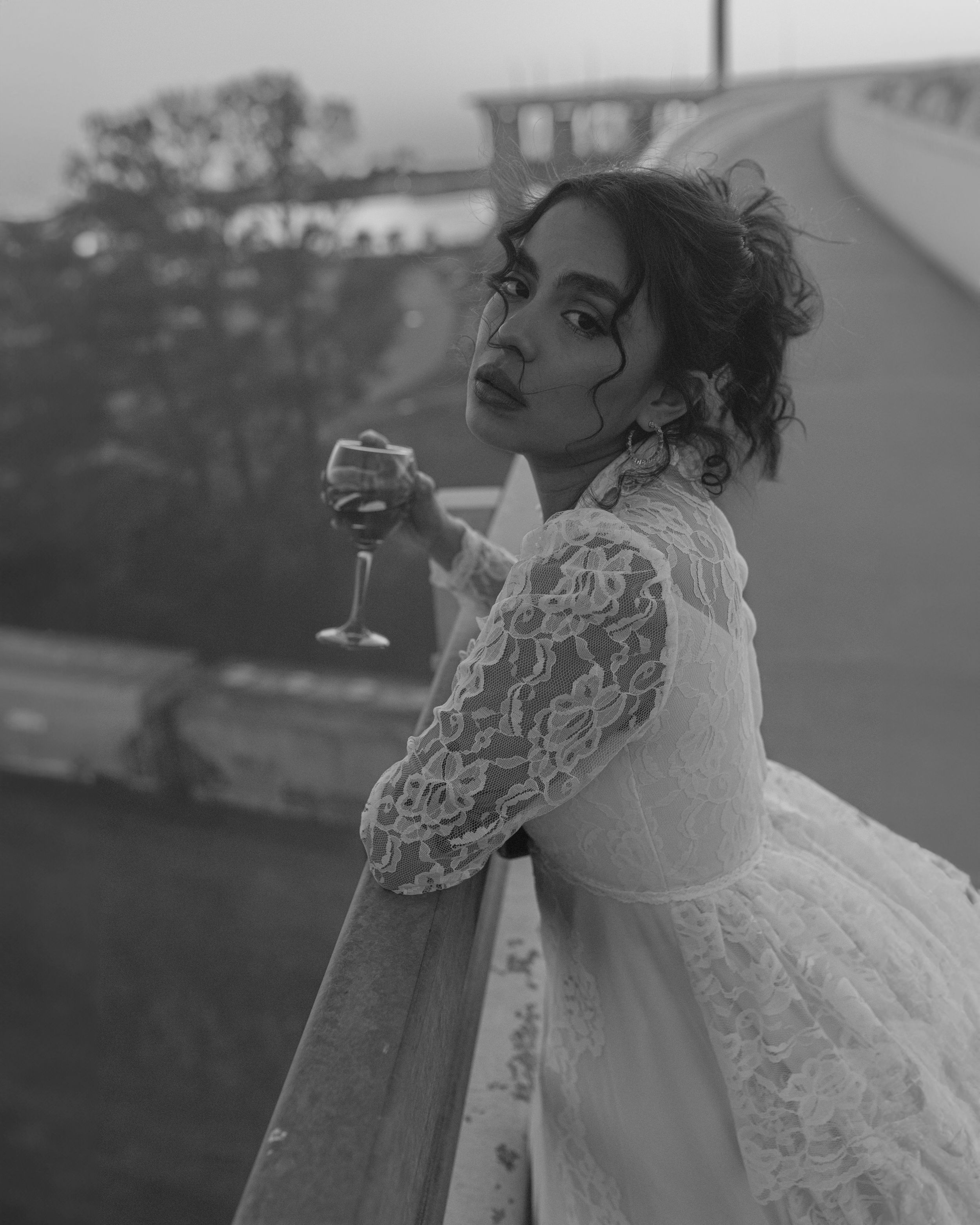 A vintage-style black and white portrait of someone in a delicate lace wedding dress holding a wine glass on a balcony.