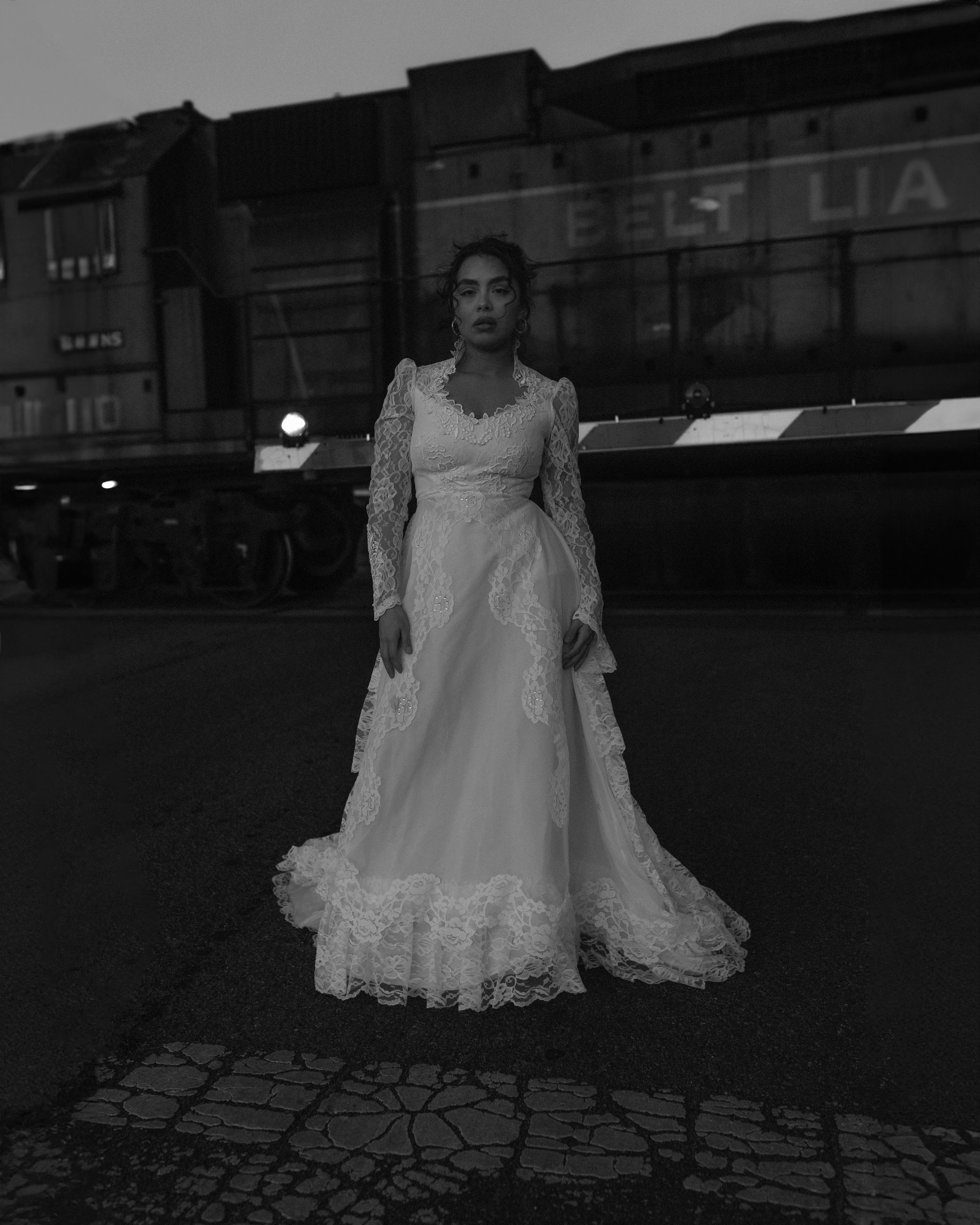A black and white photo of someone in an elegant vintage lace wedding dress standing on a city street at night.