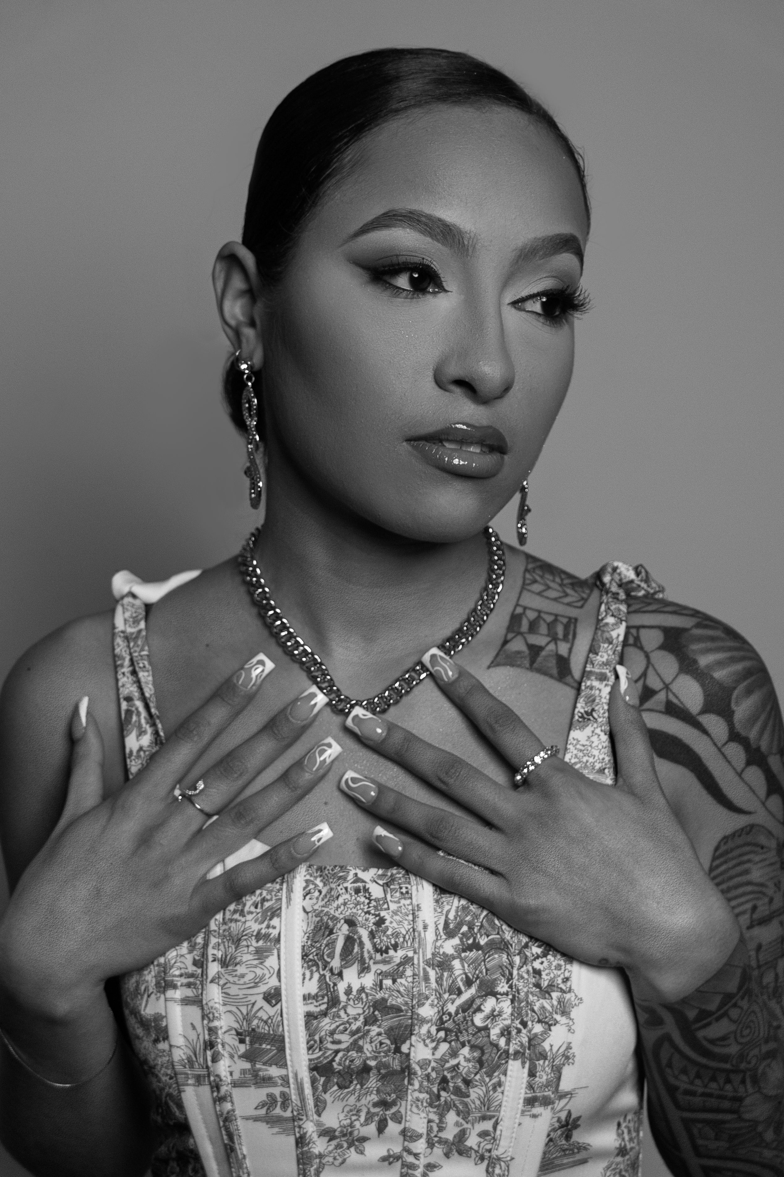 Black and white portrait showing a woman in a corset top with tattoos wearing jewelry and posing with hands crossed over chest.