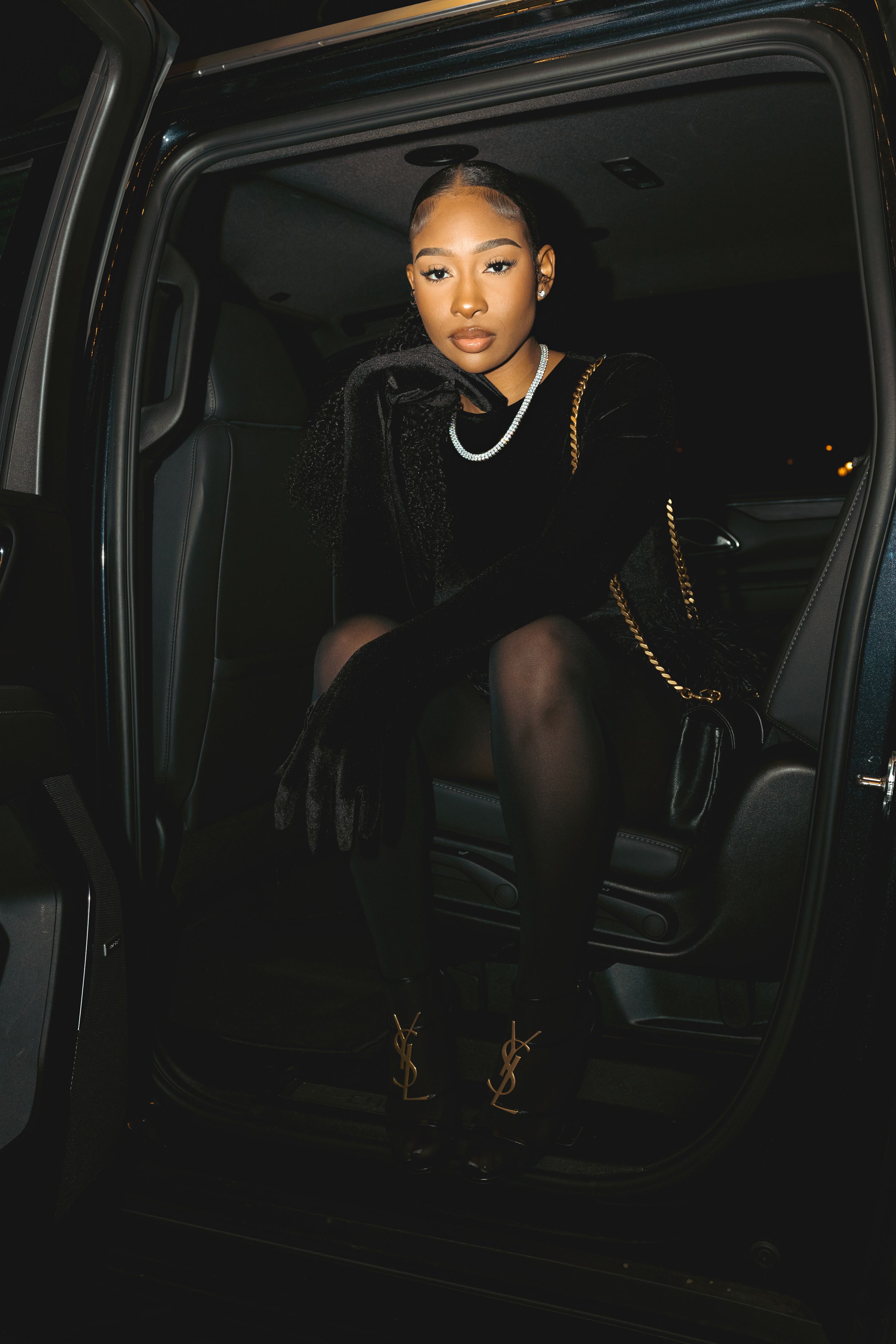 Stylish figure in black attire sitting in dark car interior with gold chain accessories and boots in moody lighting.
