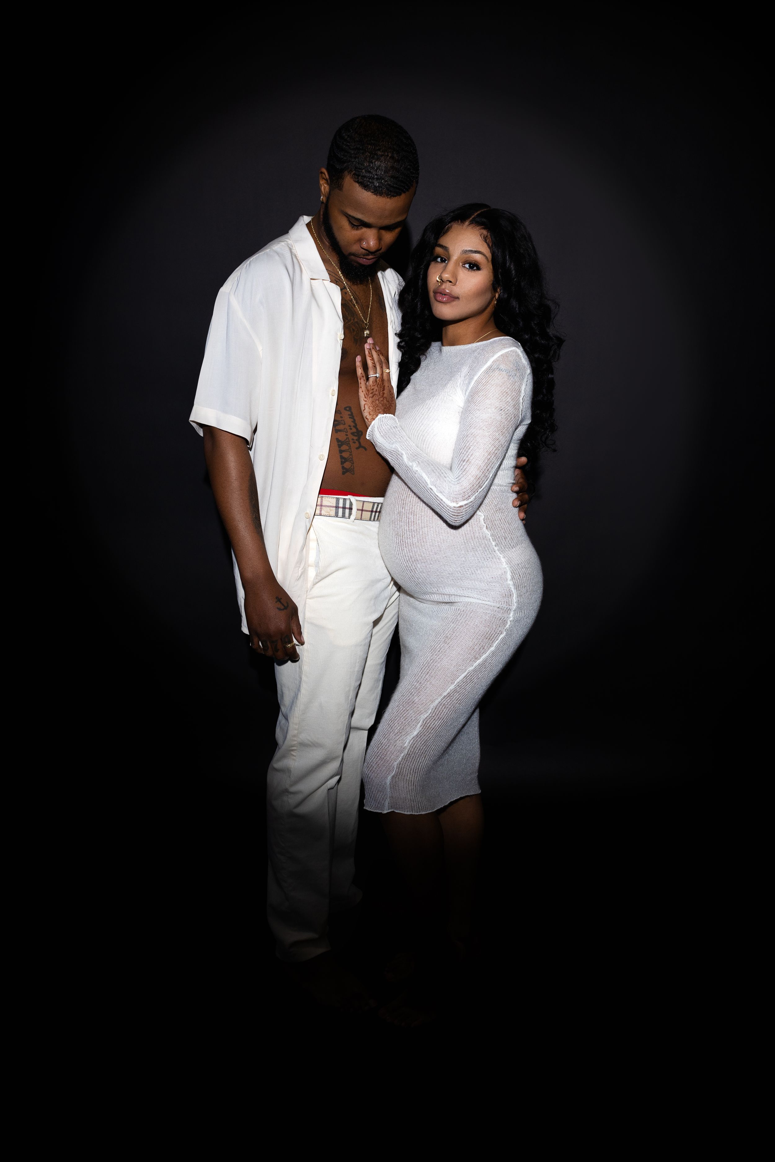 A couple poses intimately against a dark background wearing matching white outfits in a romantic photoshoot.
