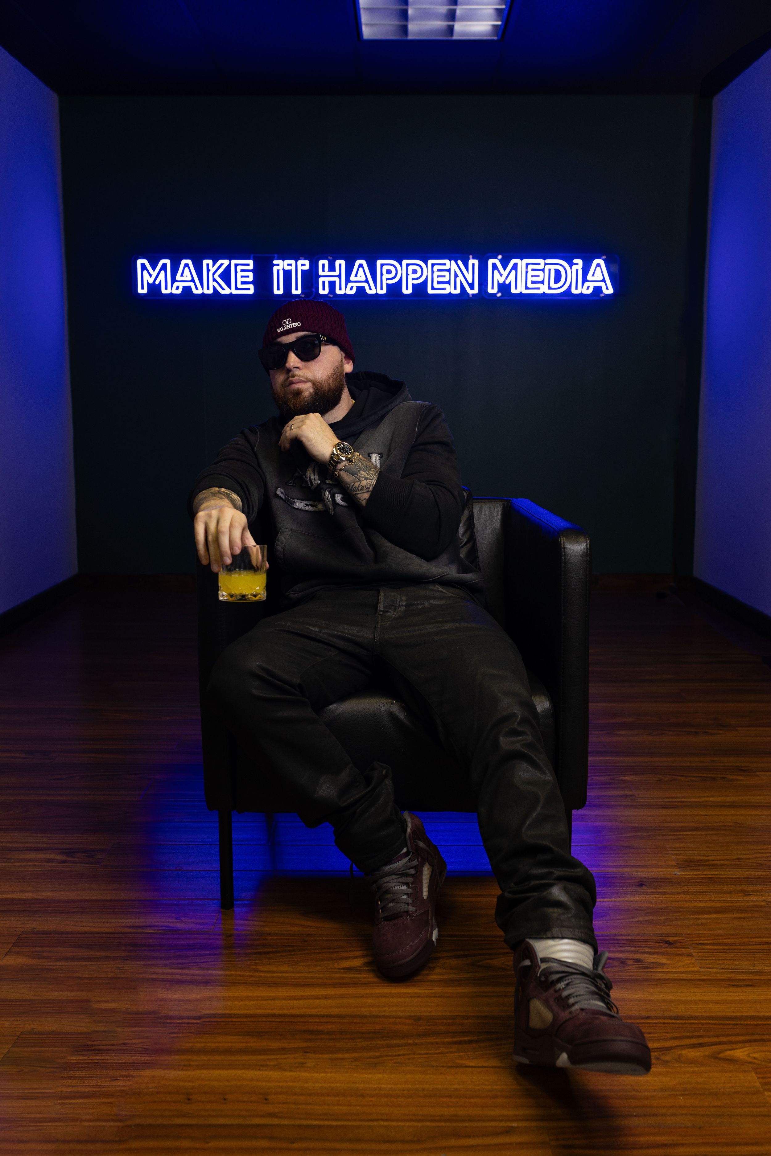 Person sitting in dark room with blue neon 'Make it Happen Media' sign wearing sunglasses with drink on armrest.