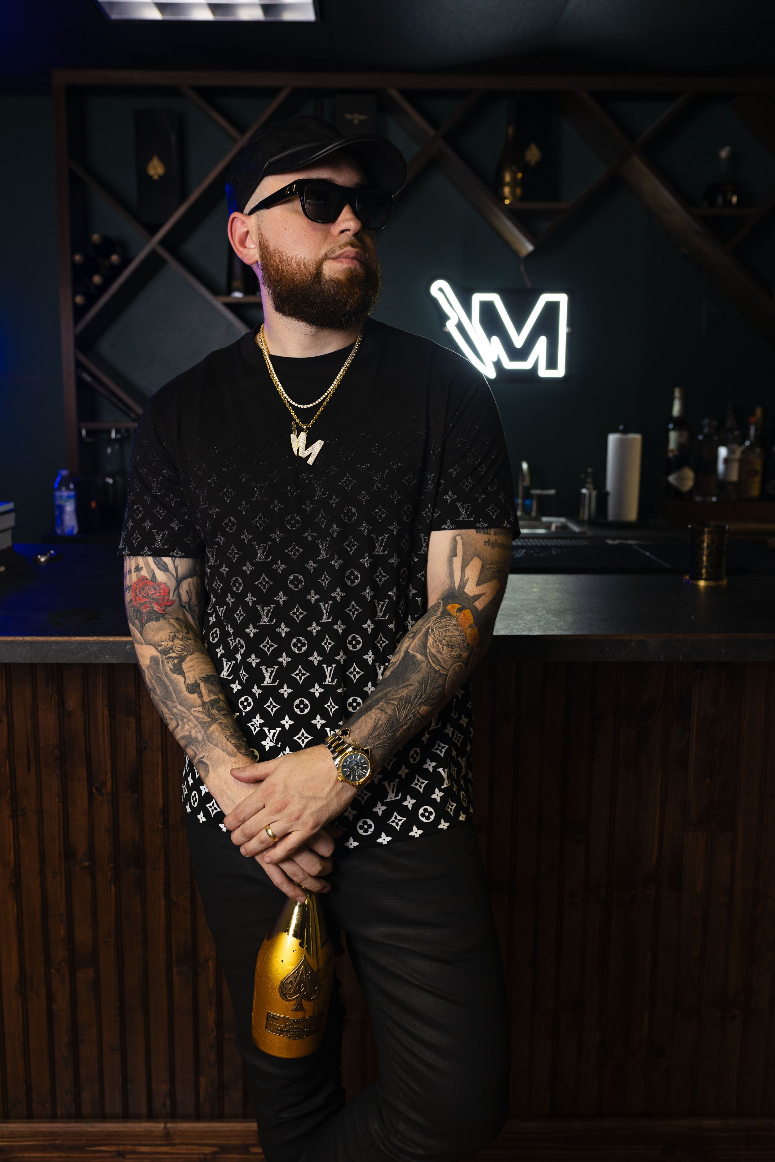 Man in sunglasses wearing black patterned t-shirt poses with bottle in dark bar with neon sign.