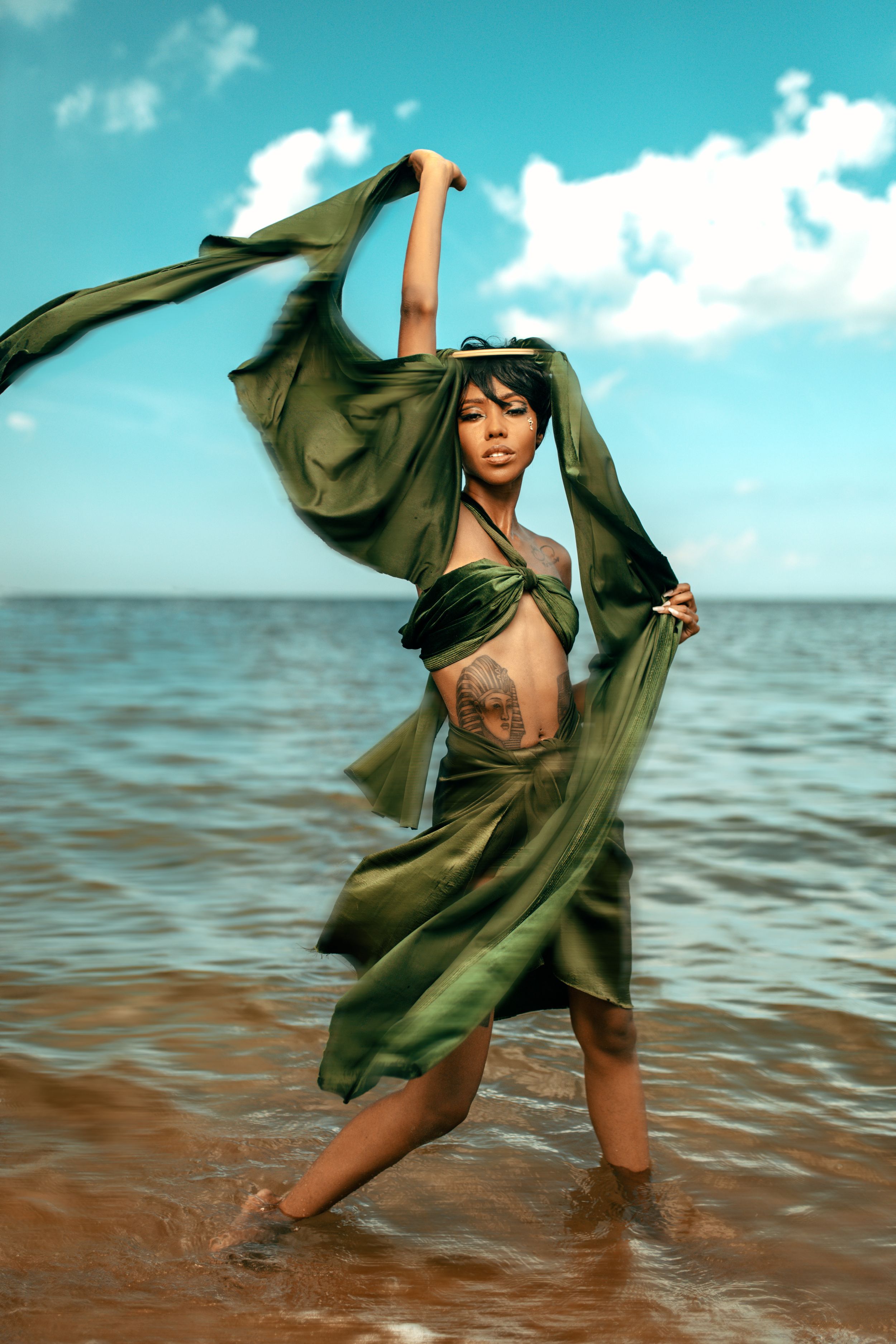 A model poses gracefully in flowing olive green fabric at the beach, with turquoise water and sky in the background.