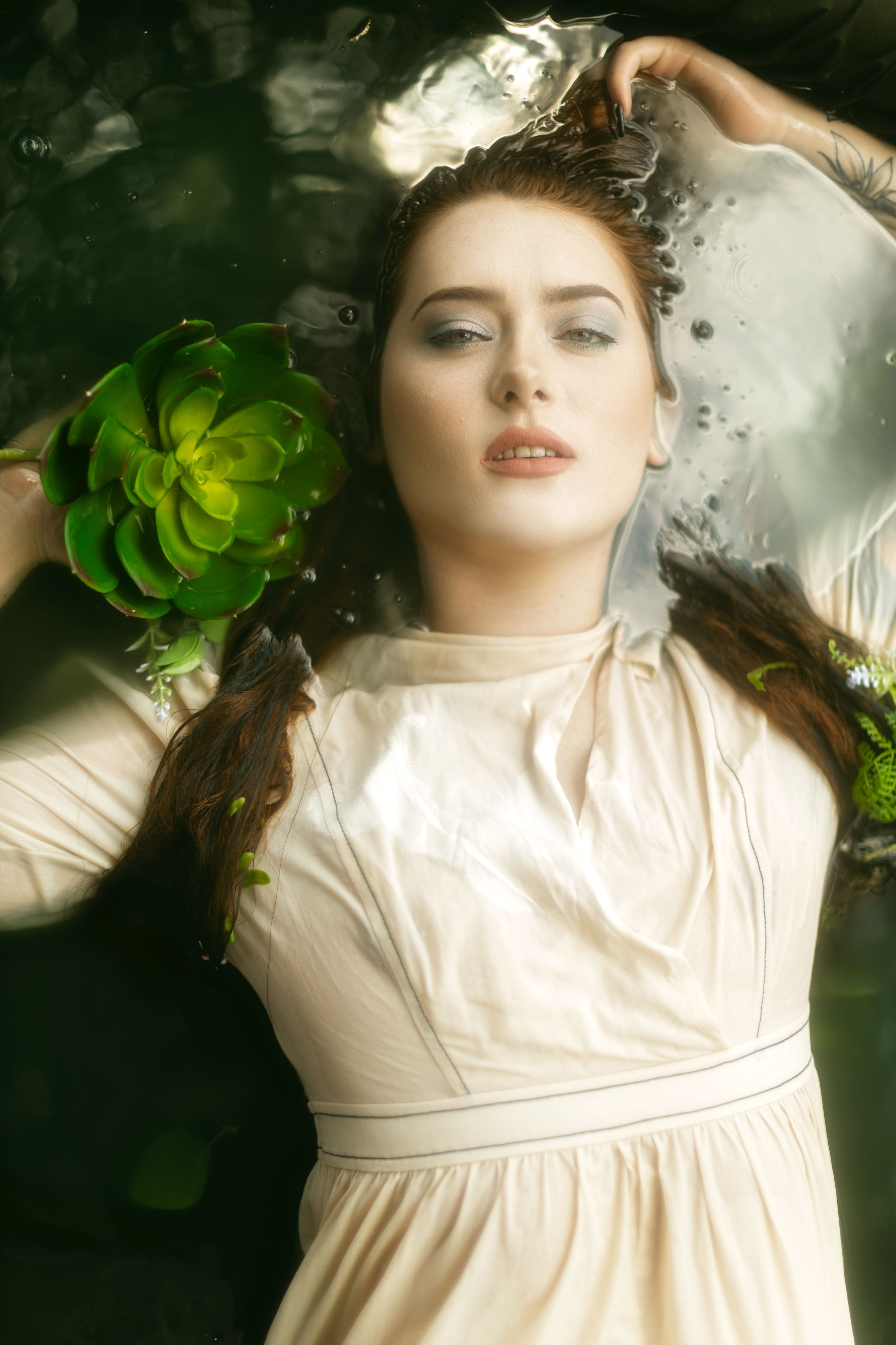 Ethereal portrait in cream dress with green foliage floating in dark water creates dreamy artistic atmosphere.