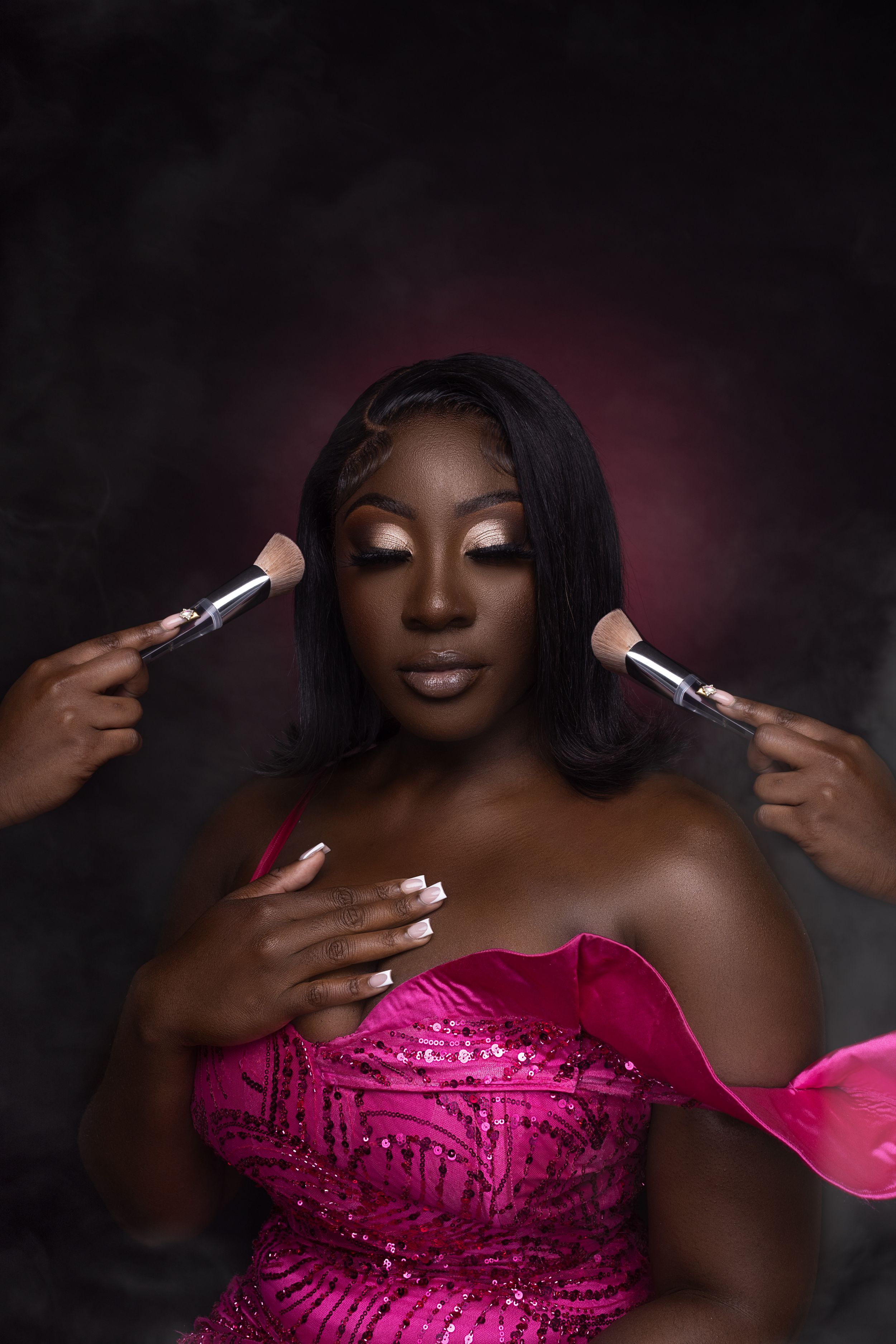 Hands holding makeup brushes apply cosmetics to person in glamorous pink dress against dark dramatic background.