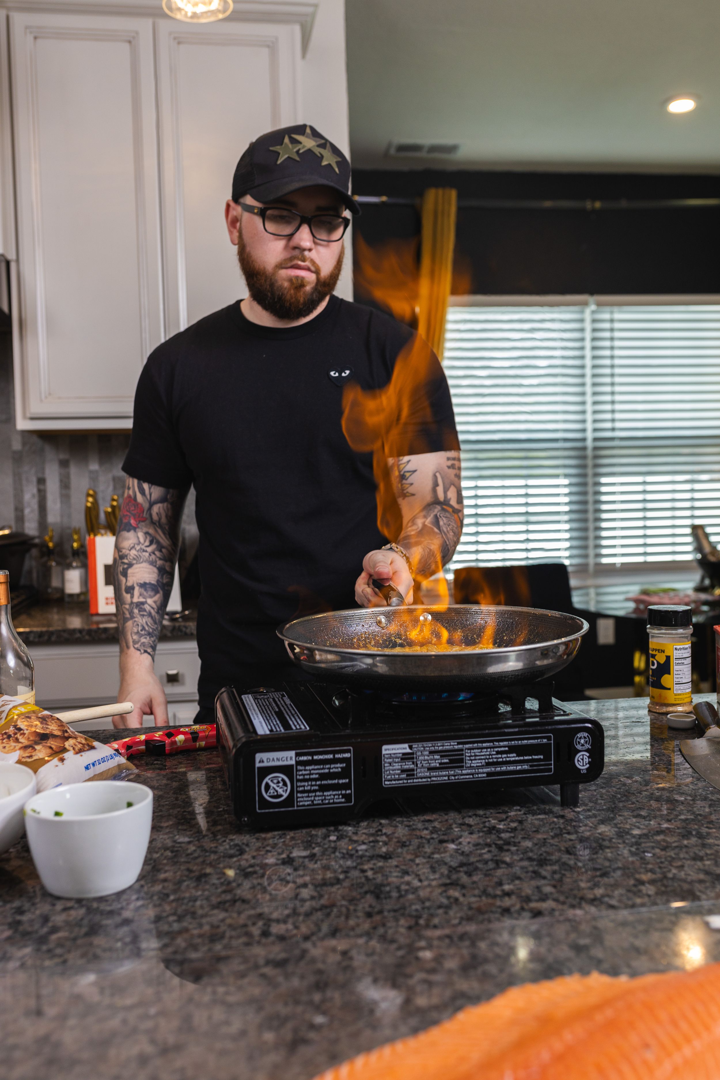 Chef cooking with flames in a pan on portable burner in modern kitchen with granite countertops.