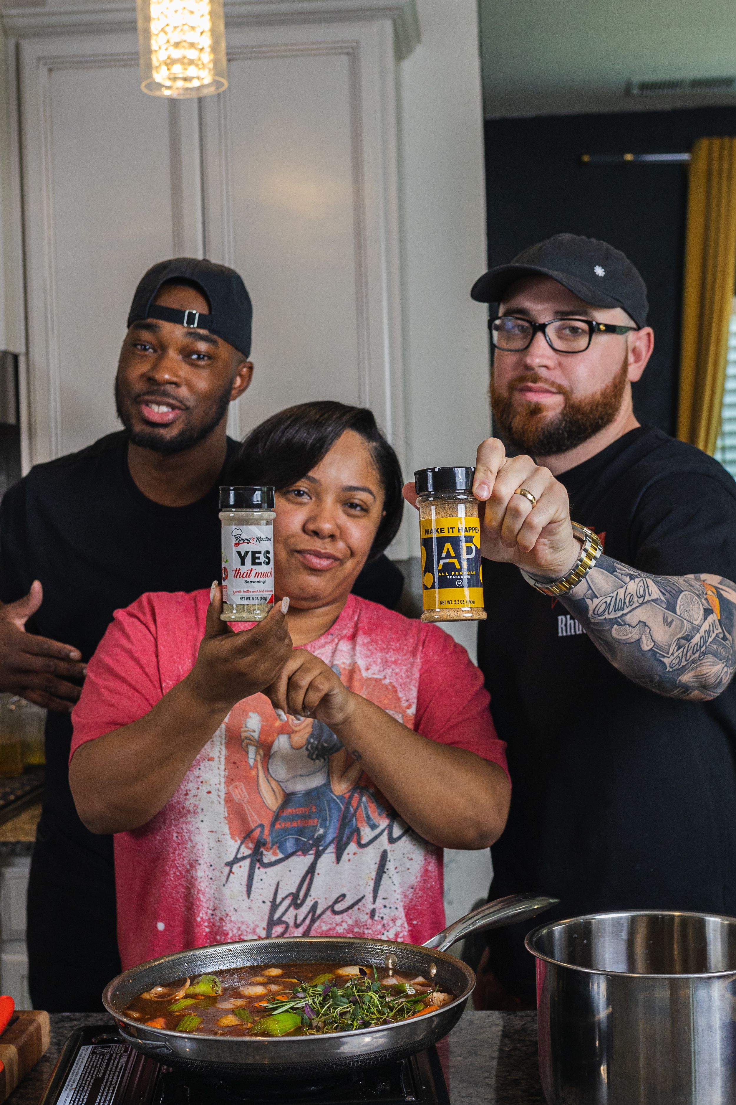 Three friends pose with cans of beer while cooking a steaming dish in a pan at home.