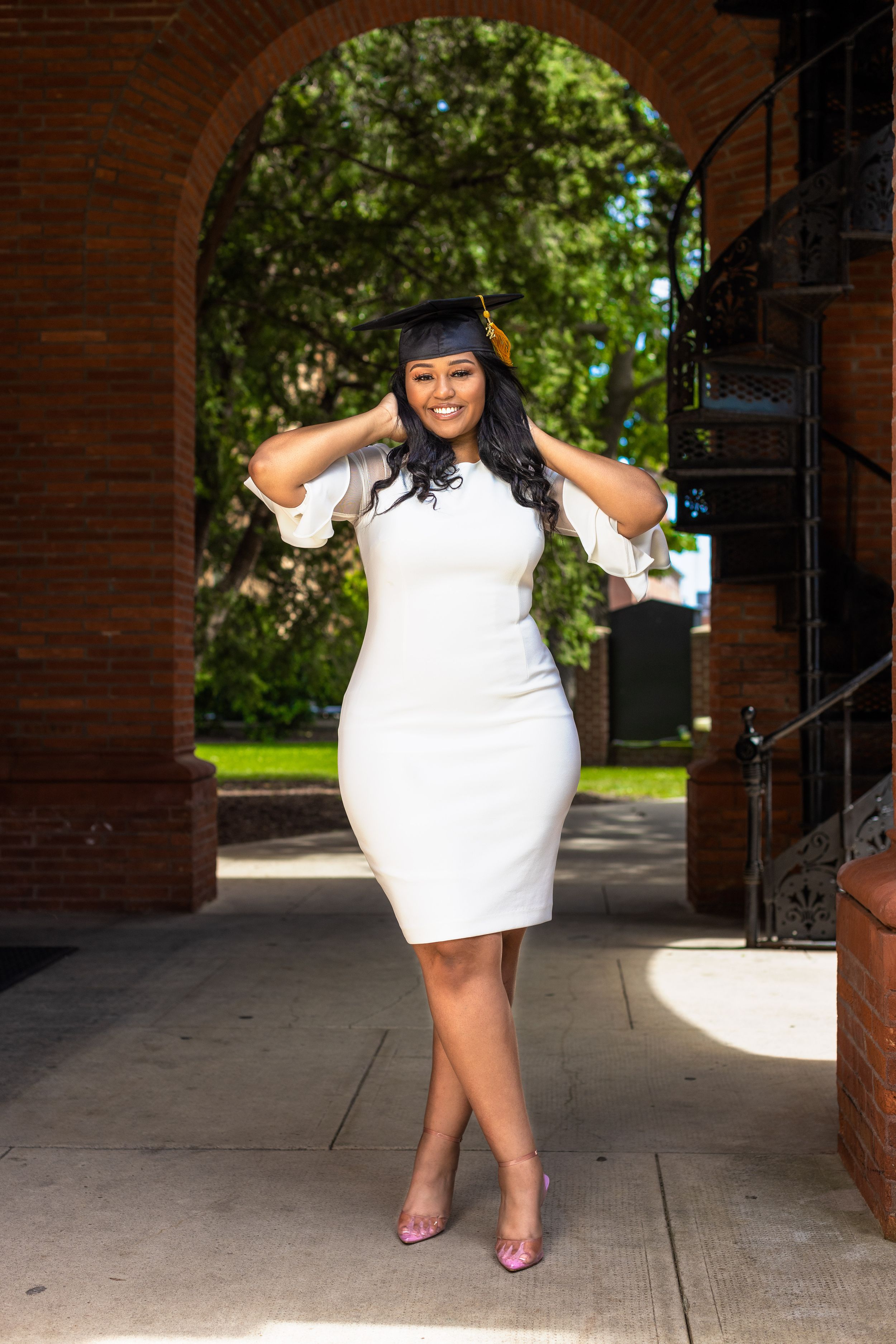 Graduate in white dress and graduation cap poses under brick archway flanked by trees and greenery.