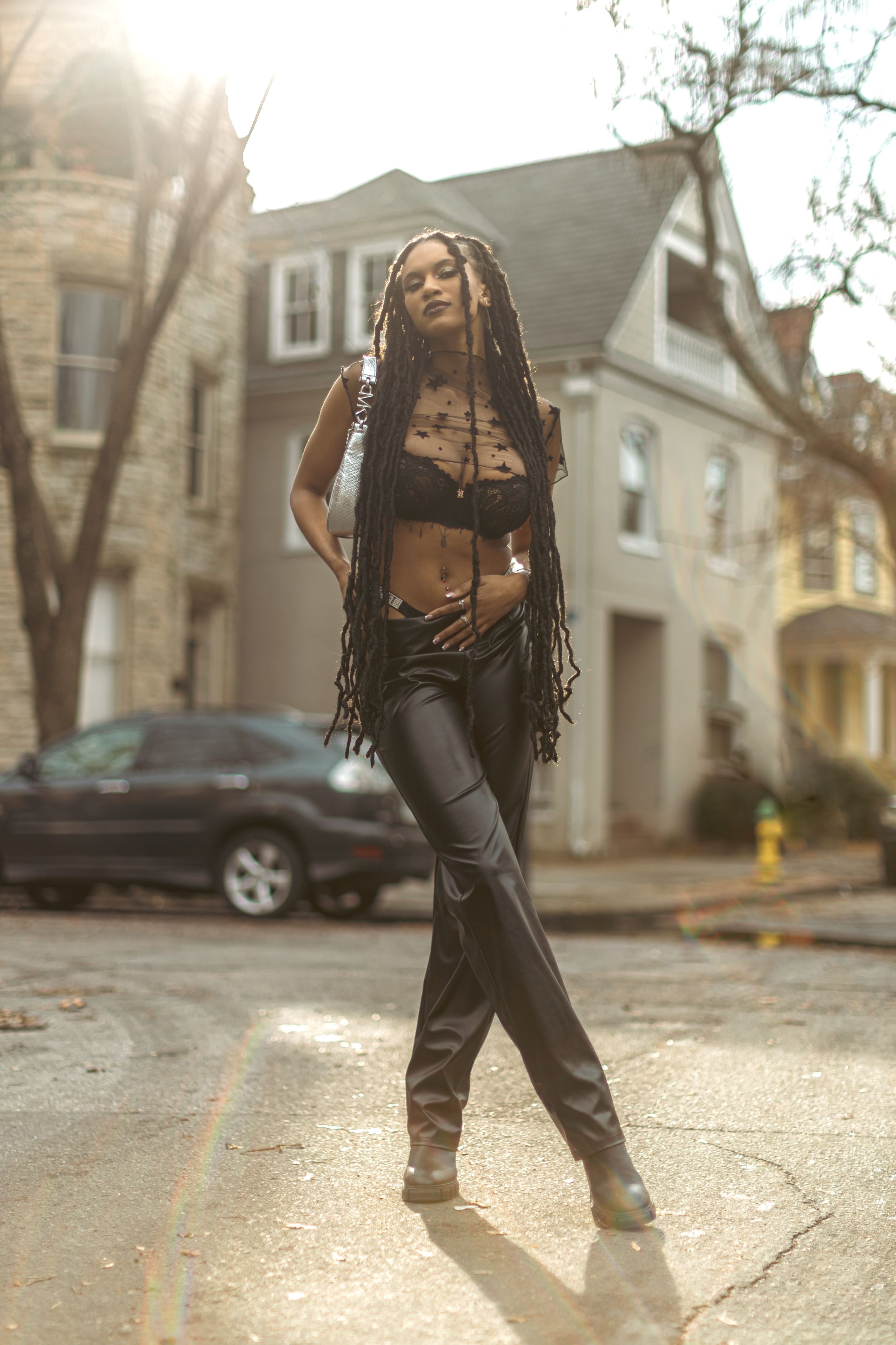 A fashion model in black leather pants and crop top poses on a city street with long braided hair during golden hour.