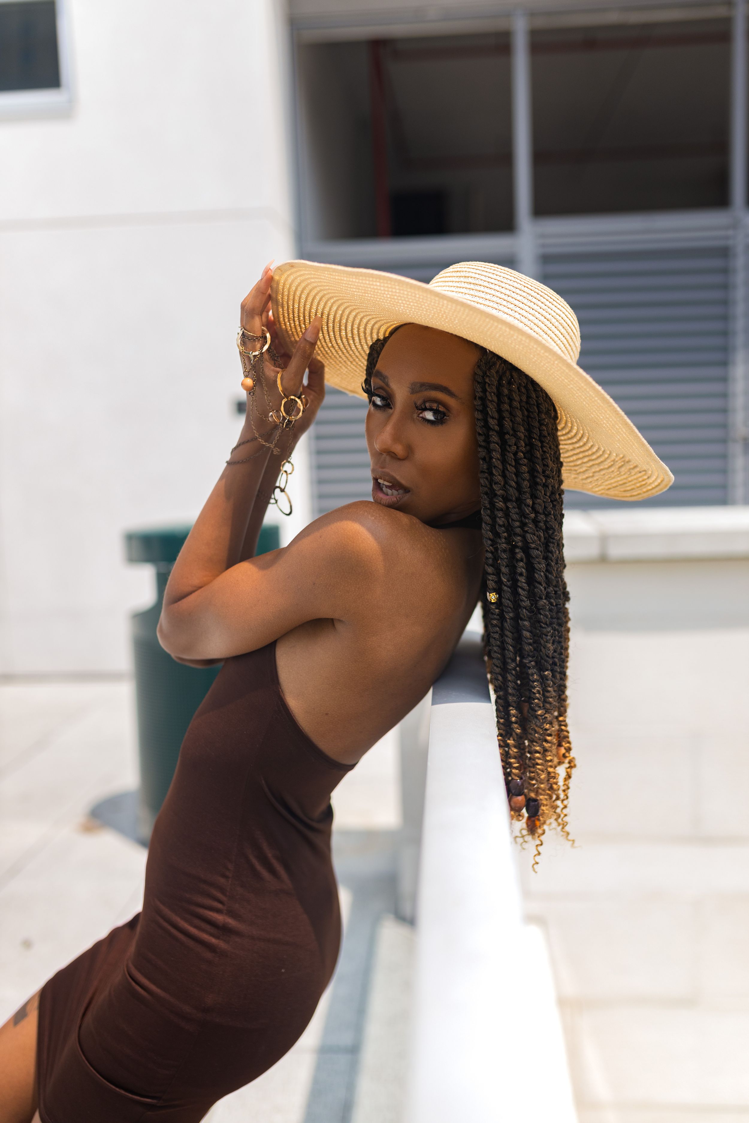 A stylish summer portrait with a straw hat and long braided hair against a white wall backdrop.