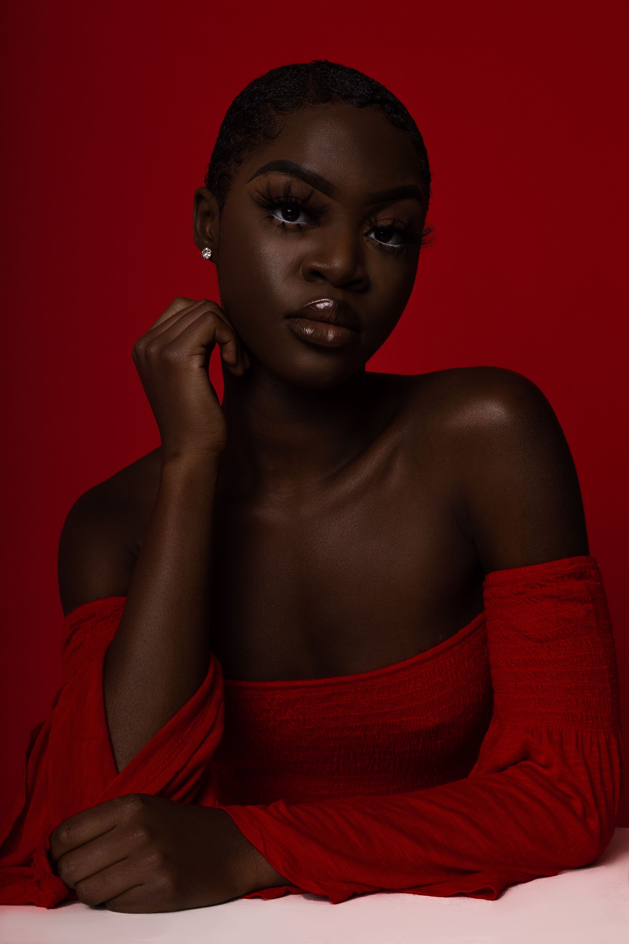 A striking fashion portrait against a deep red background features elegant off-shoulder red clothing and radiant lighting.