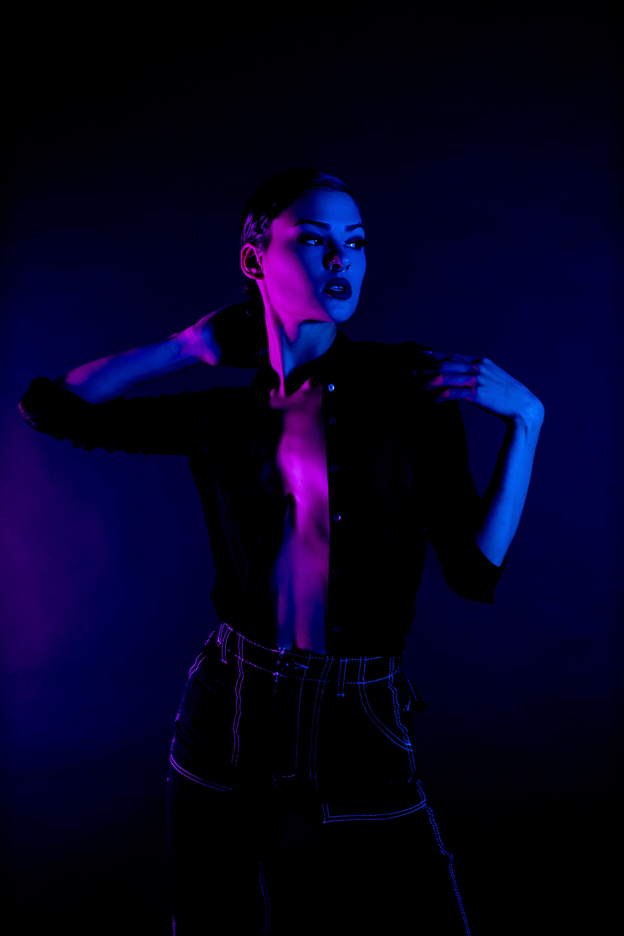 Artistic portrait in neon lighting with blue and pink tones showing dramatic pose against dark background.