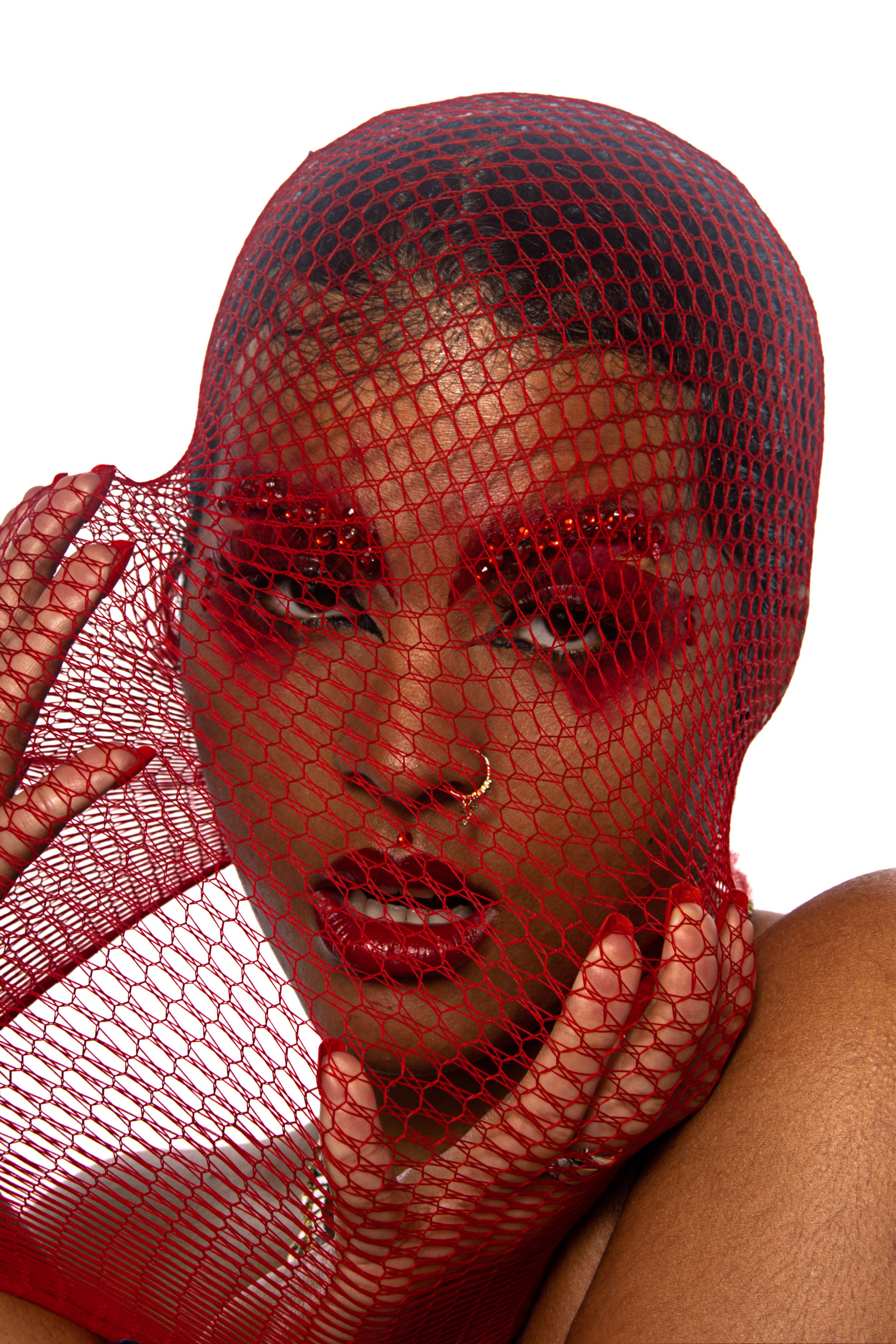 Artistic fashion portrait with model wearing red fishnet veil and bright red lipstick against white background.