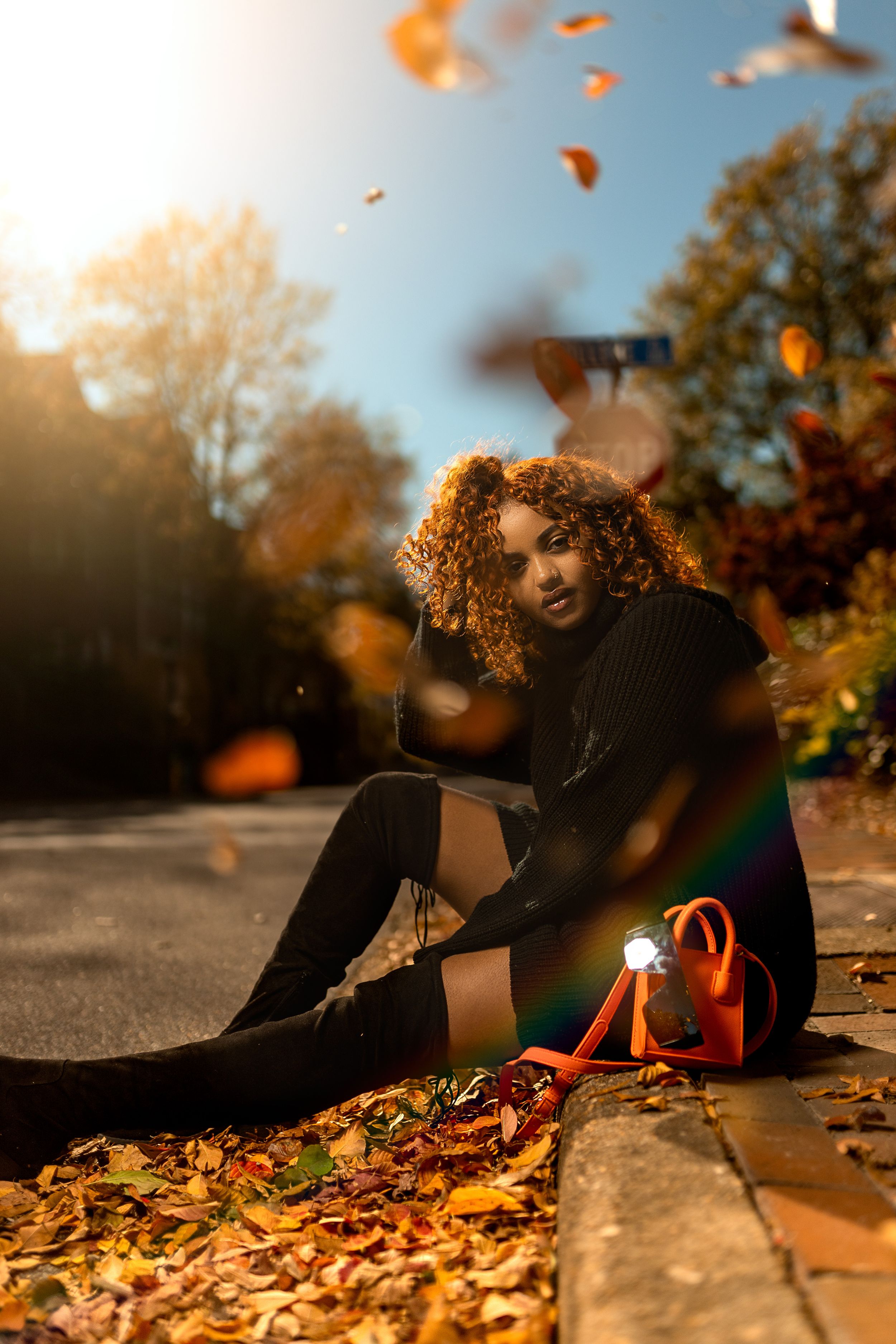 Someone sits on ground surrounded by autumn leaves during golden hour with orange foliage falling in the sunlight.