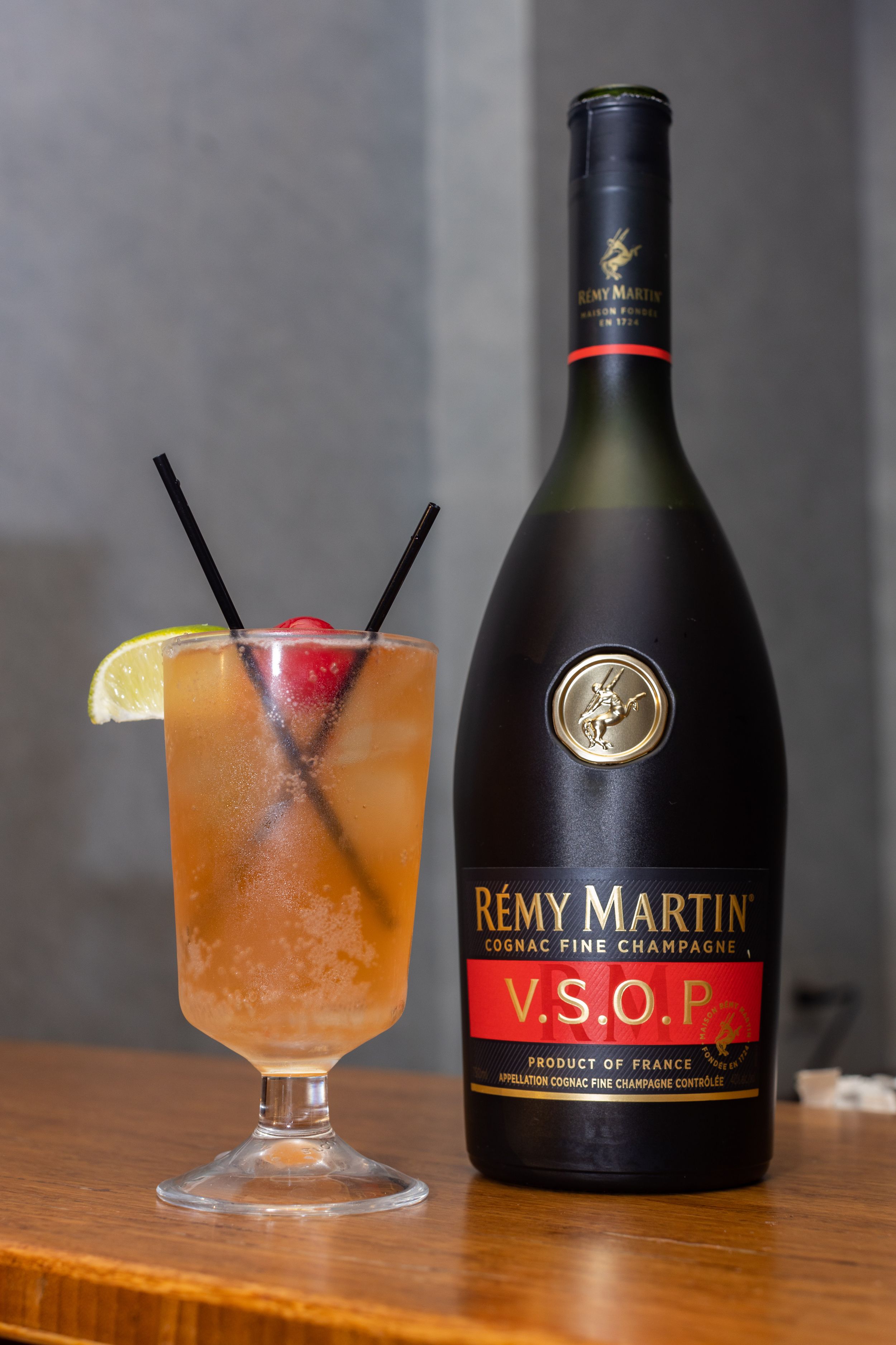 A bottle of Remy Martin VSOP cognac next to a tropical cocktail garnished with lemon and cherry on a wooden surface.