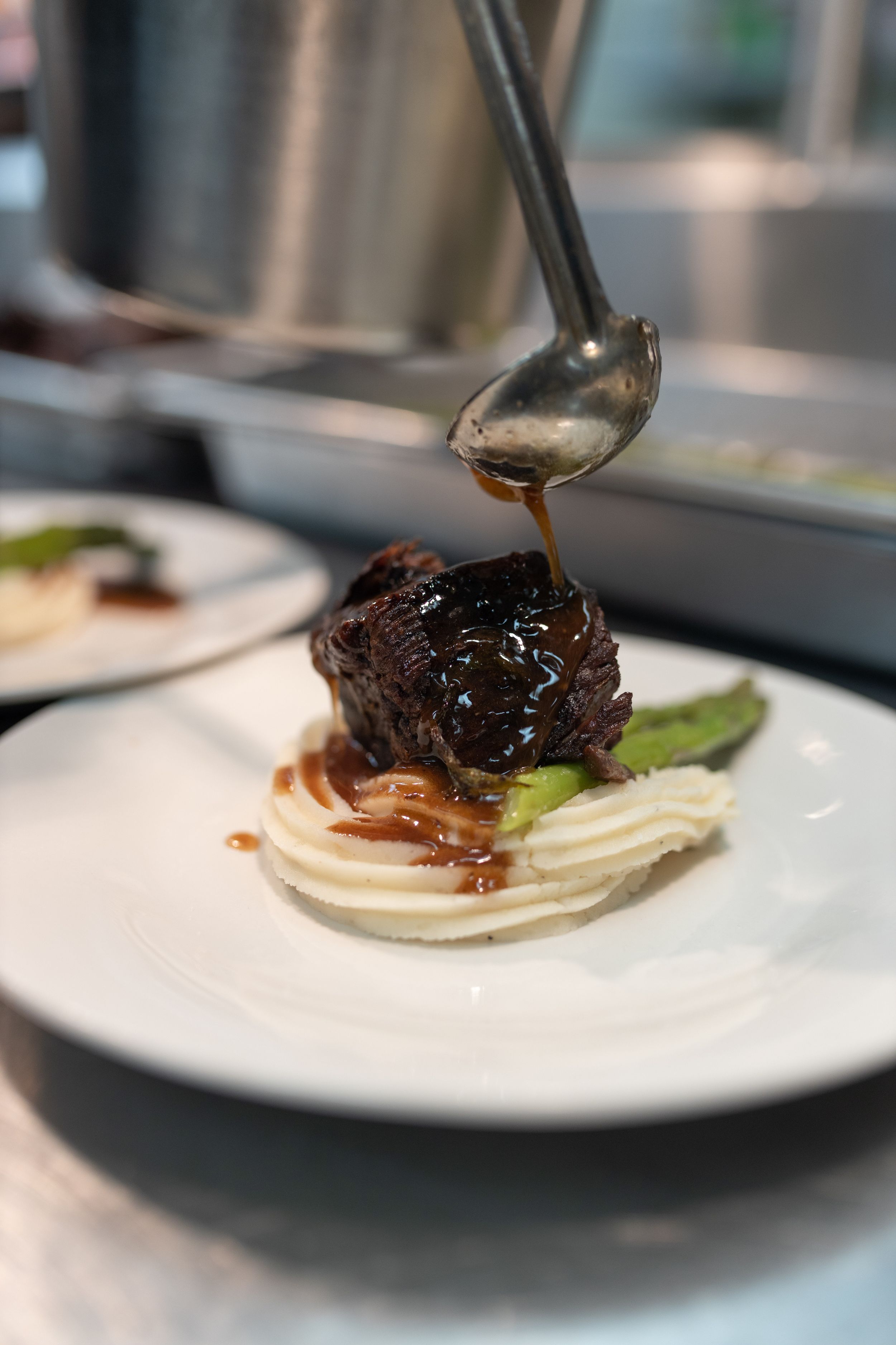 A braised short rib dish being garnished with a drizzle of sauce over mashed potatoes and asparagus on a white plate.