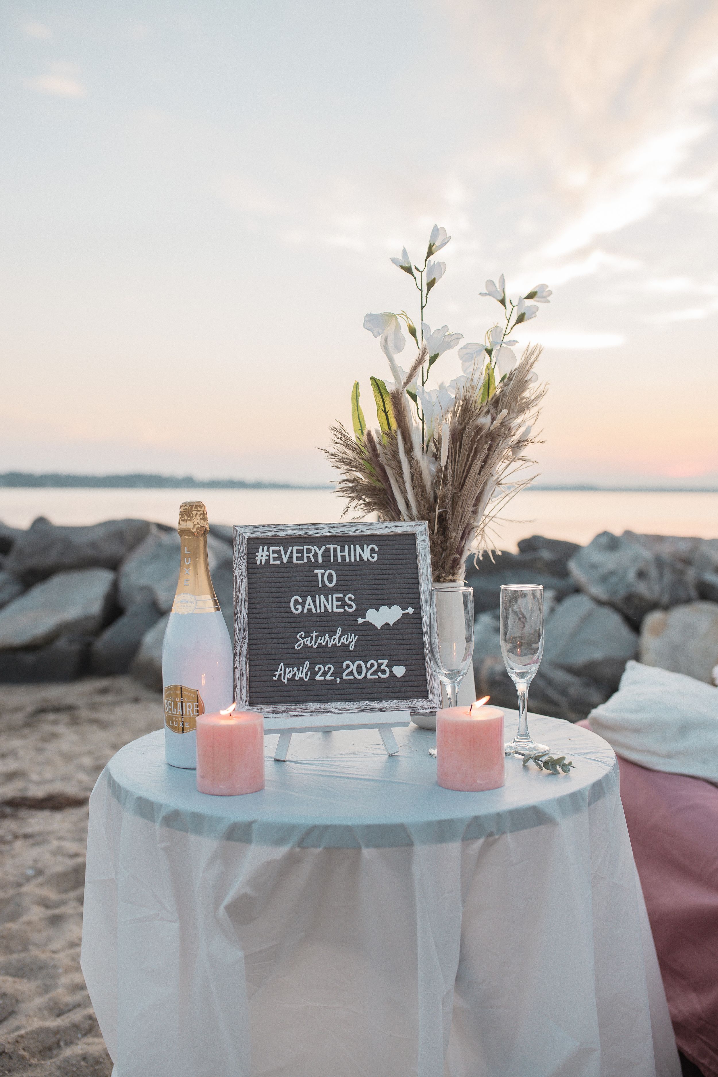 Romantic beach proposal setup with champagne, pink candles, pampas grass and a chalkboard sign at sunset by the water.