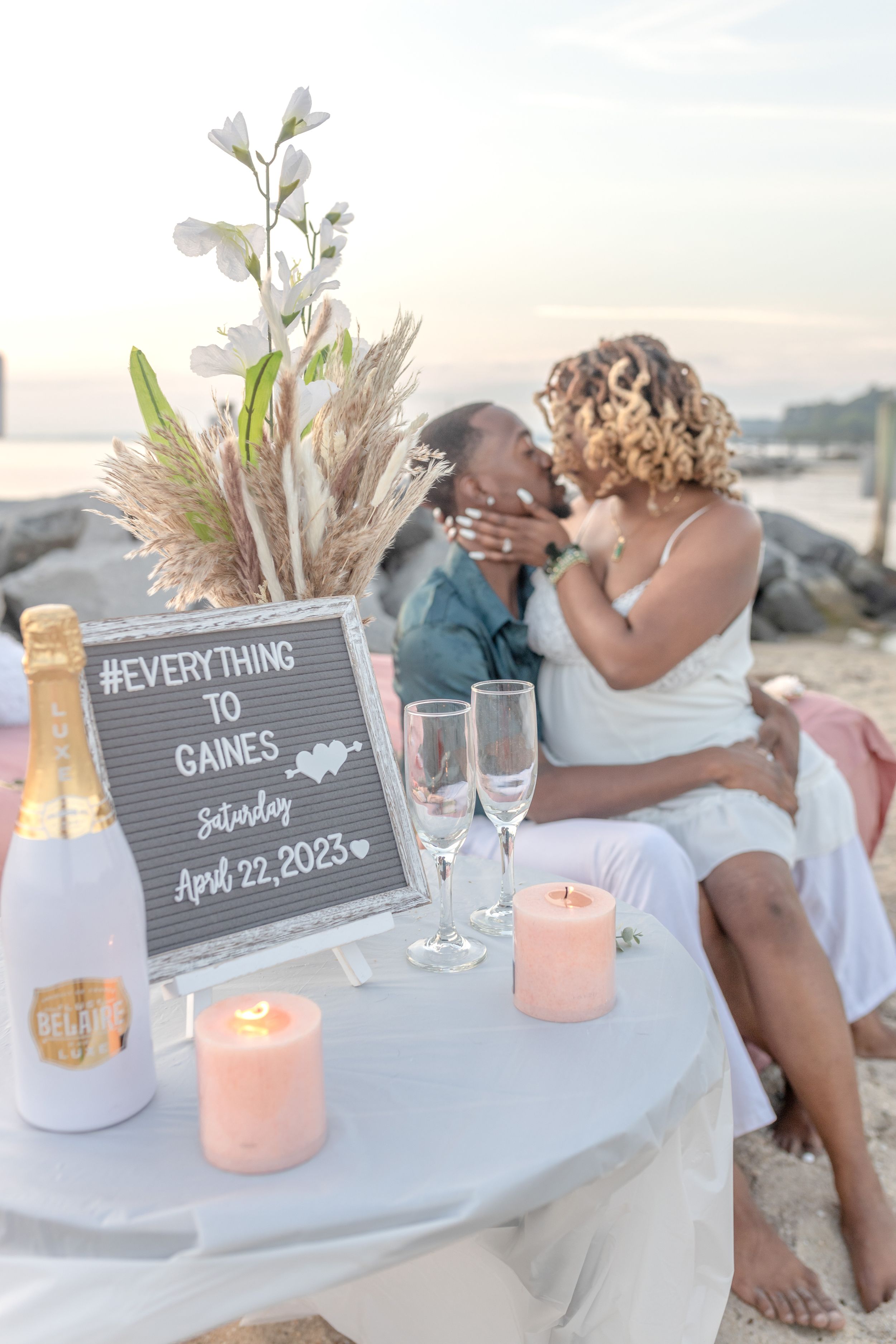 Romantic beachside engagement setting with champagne glasses, candles and signboard displaying wedding date against ocean view.