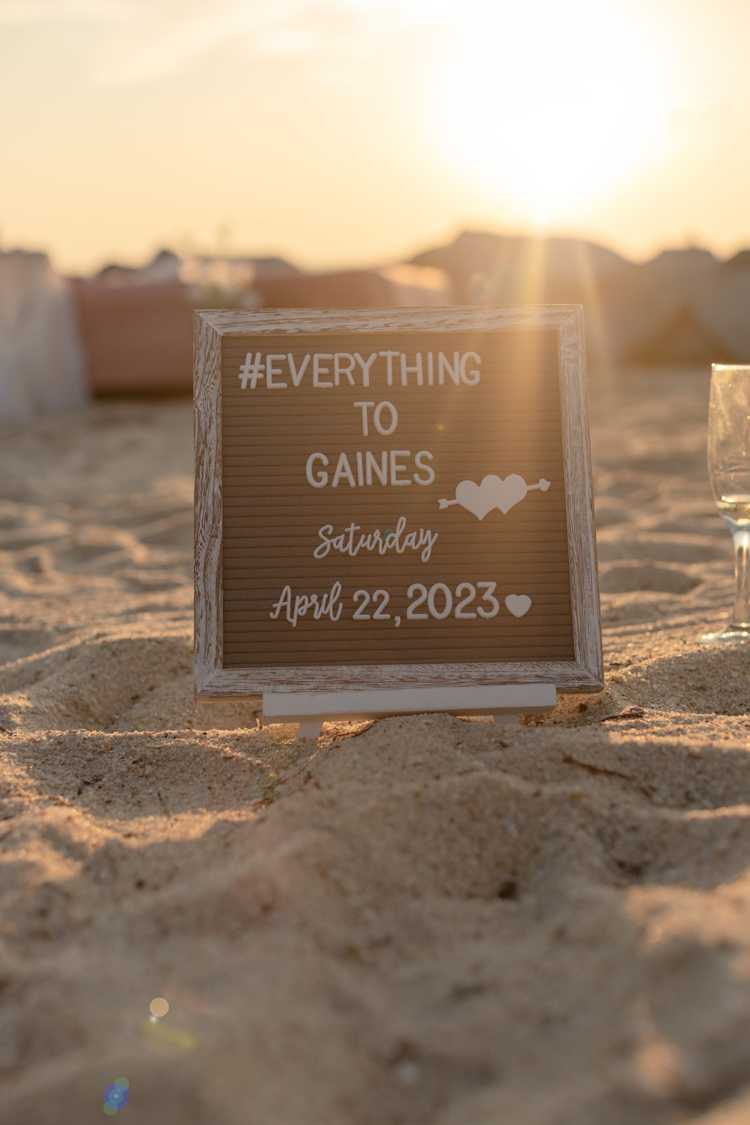 A wooden wedding sign reading '#EVERYTHING TO GAINES' sits in beach sand at sunset with April 22, 2023 date.