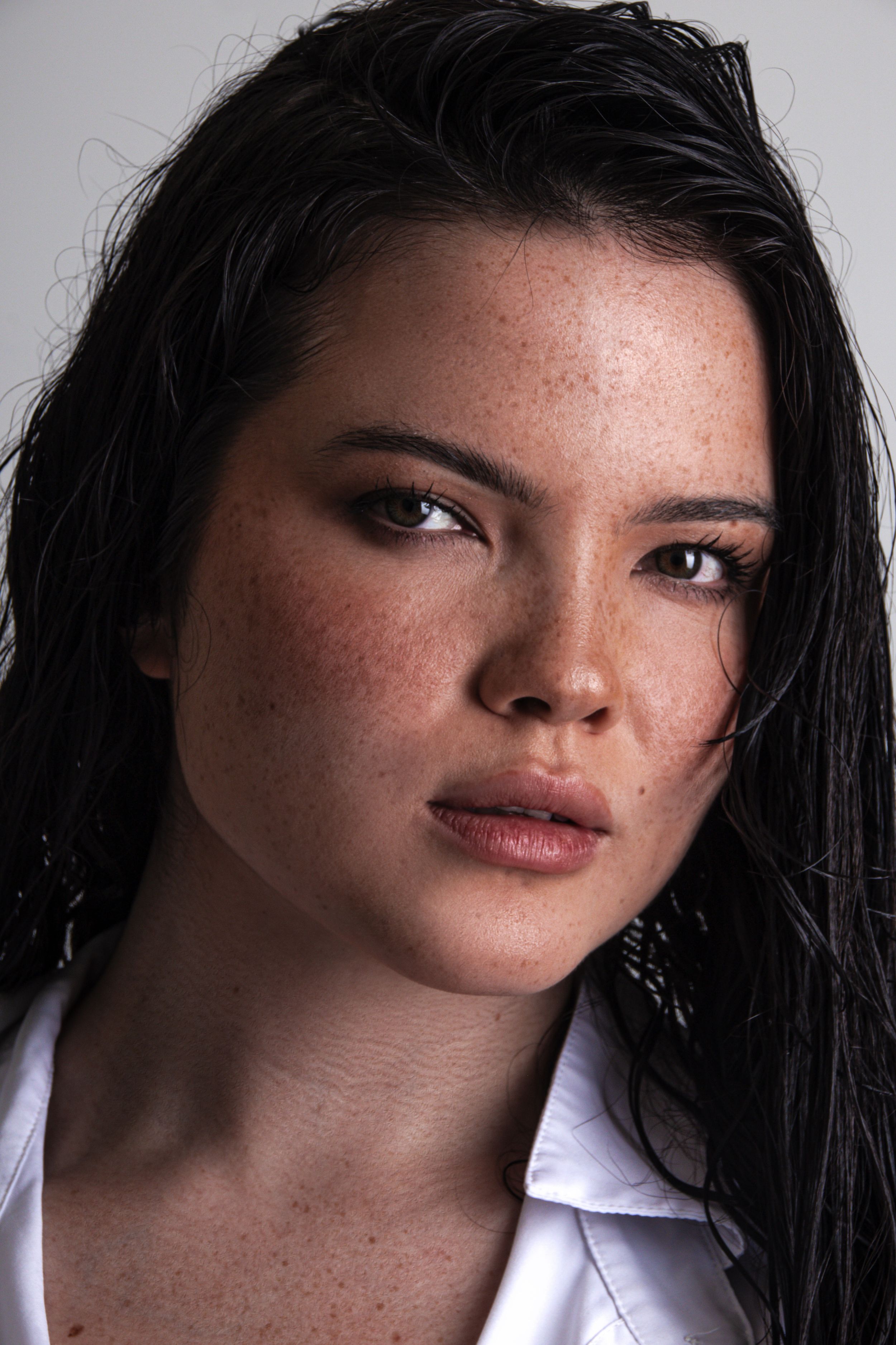 A closeup portrait of a young woman with long dark hair wearing a white shirt against a neutral background.