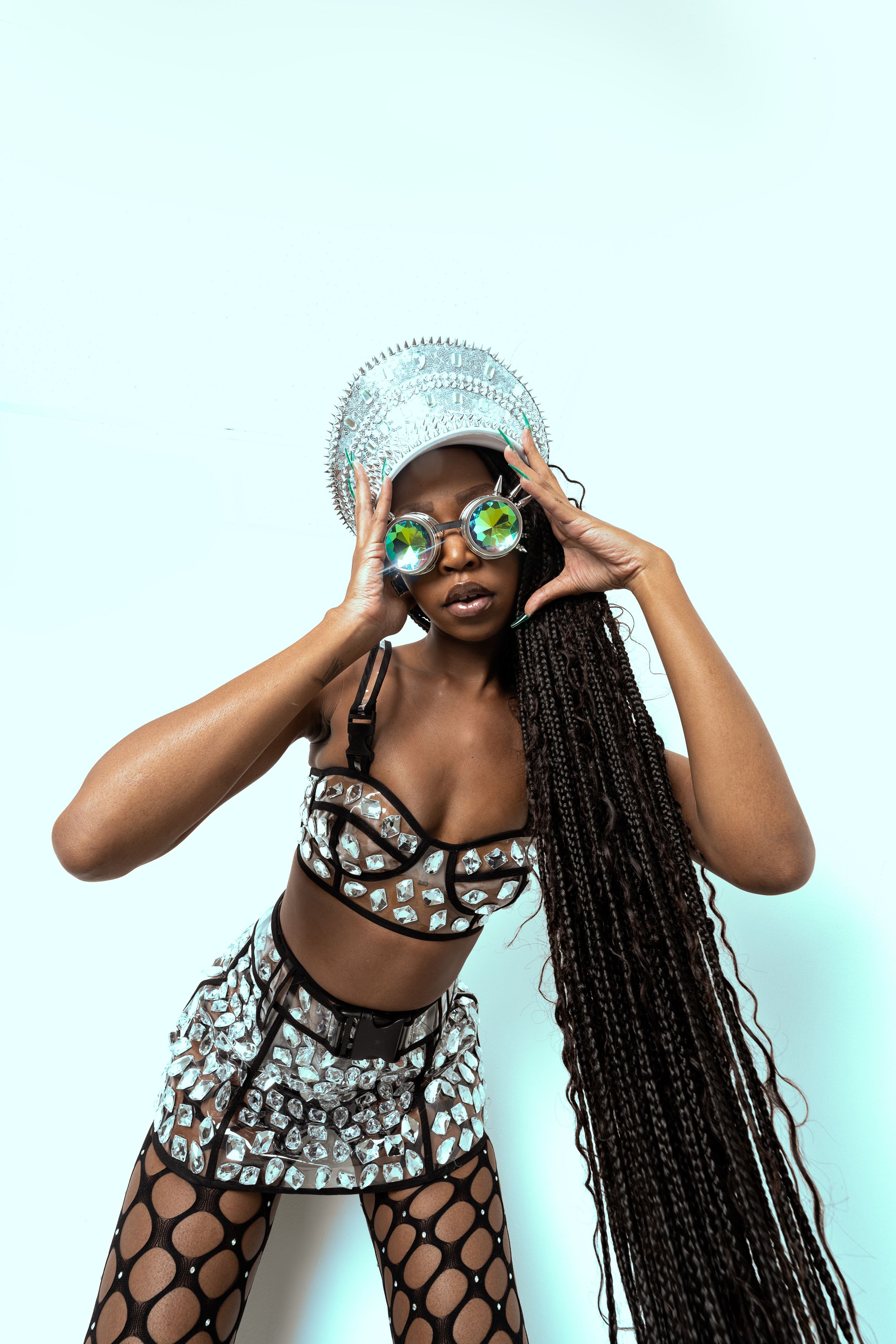 Fashion model wearing sparkly headpiece, kaleidoscope glasses and a silver embellished outfit poses against light background.