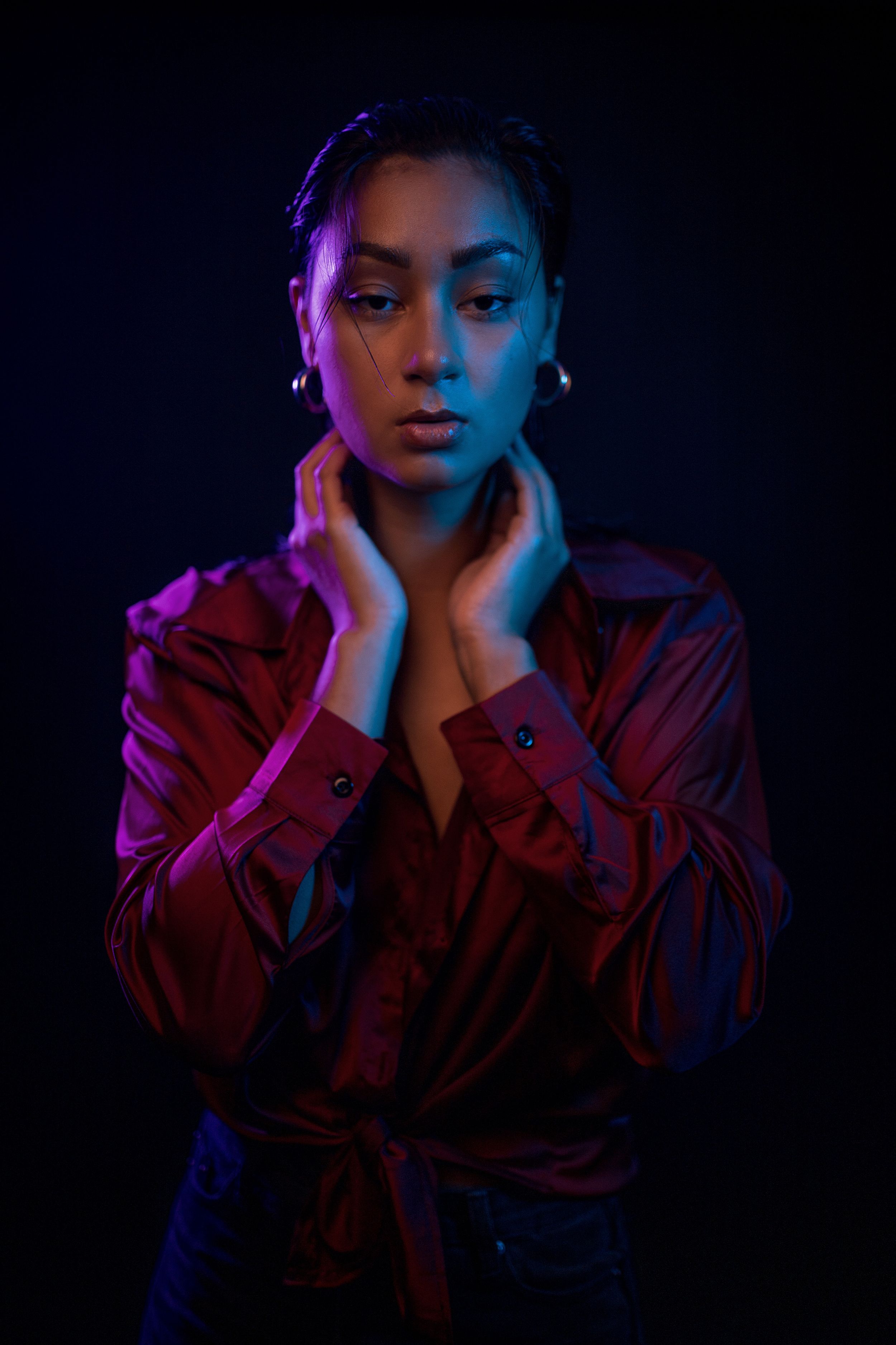 Dramatic portrait with pink and blue lighting against dark background showing figure in red jacket with hands near neck.