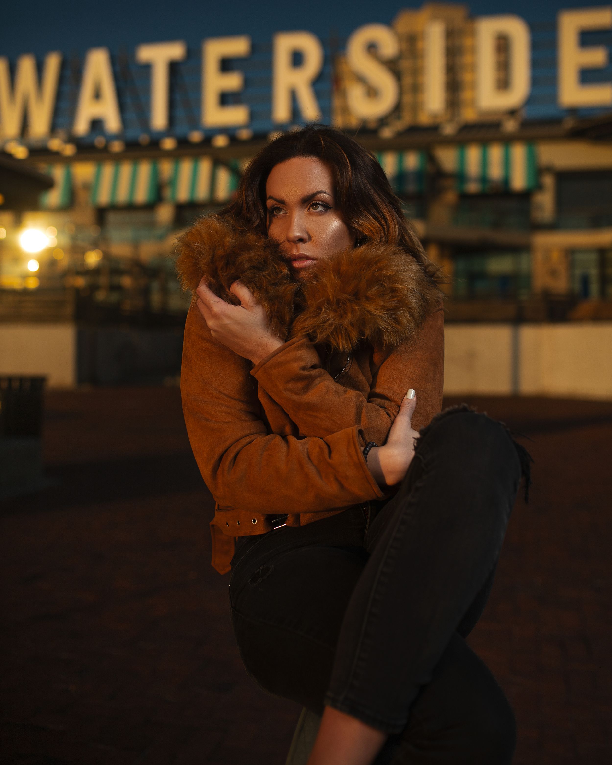 Person in brown fur-trimmed coat poses against illuminated Waterside signage at night.