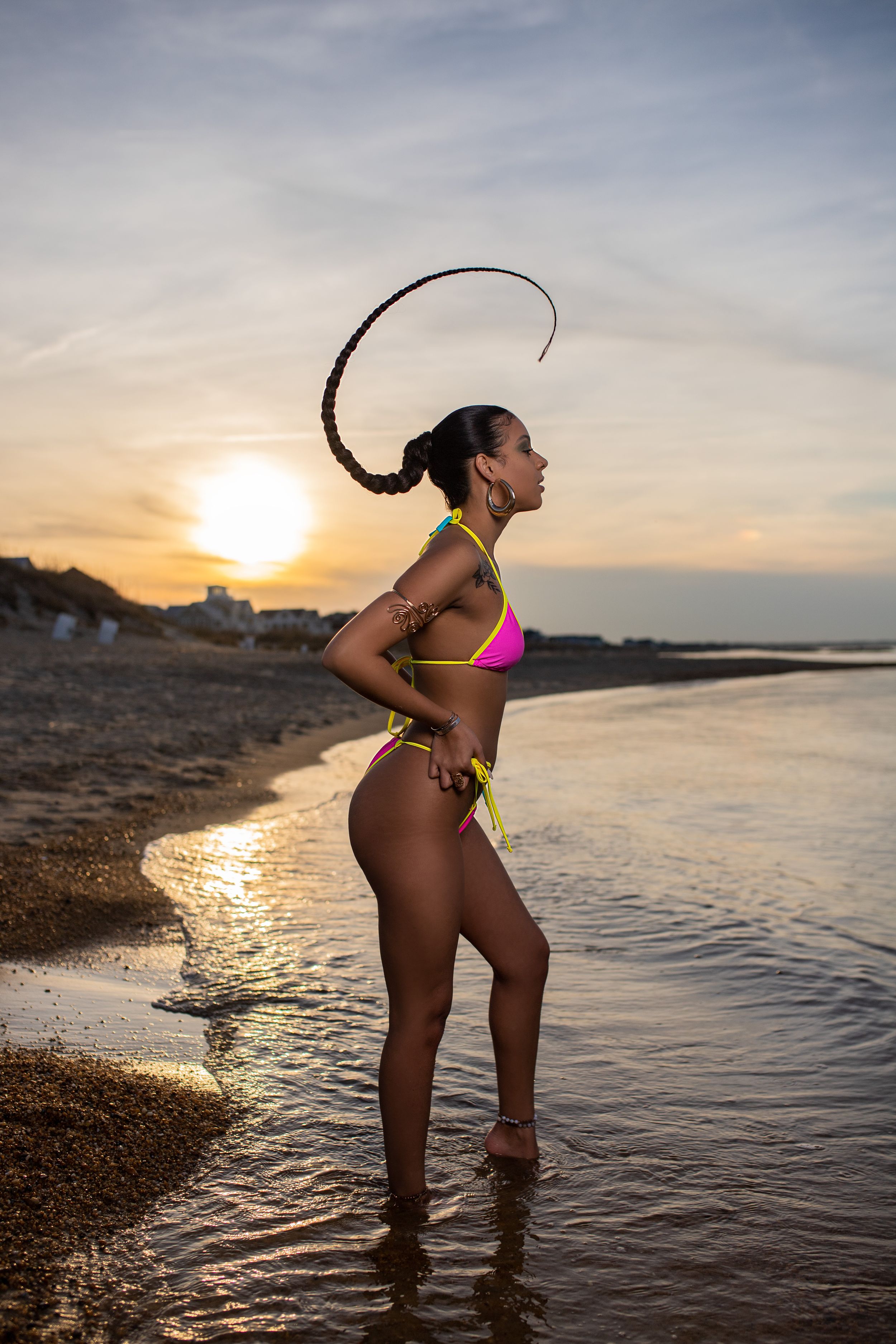Woman in pink bikini stands in shallow ocean water during sunset with hair flipping back in silhouette.