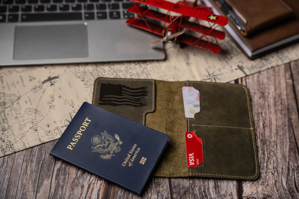 A US passport and green leather wallet lie on a wooden desk next to a laptop and red toy airplane.