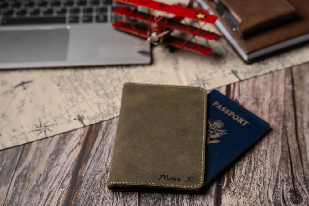 Travel documents including passport and leather cases lay near a laptop with decorative red airplane on wooden surface.
