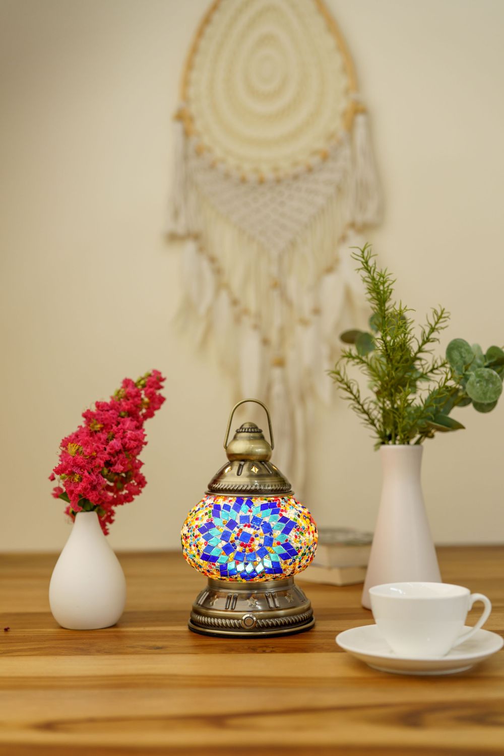 Turkish mosaic lamp on wooden table with dreamcatcher, red flowers and teacup against beige wall.