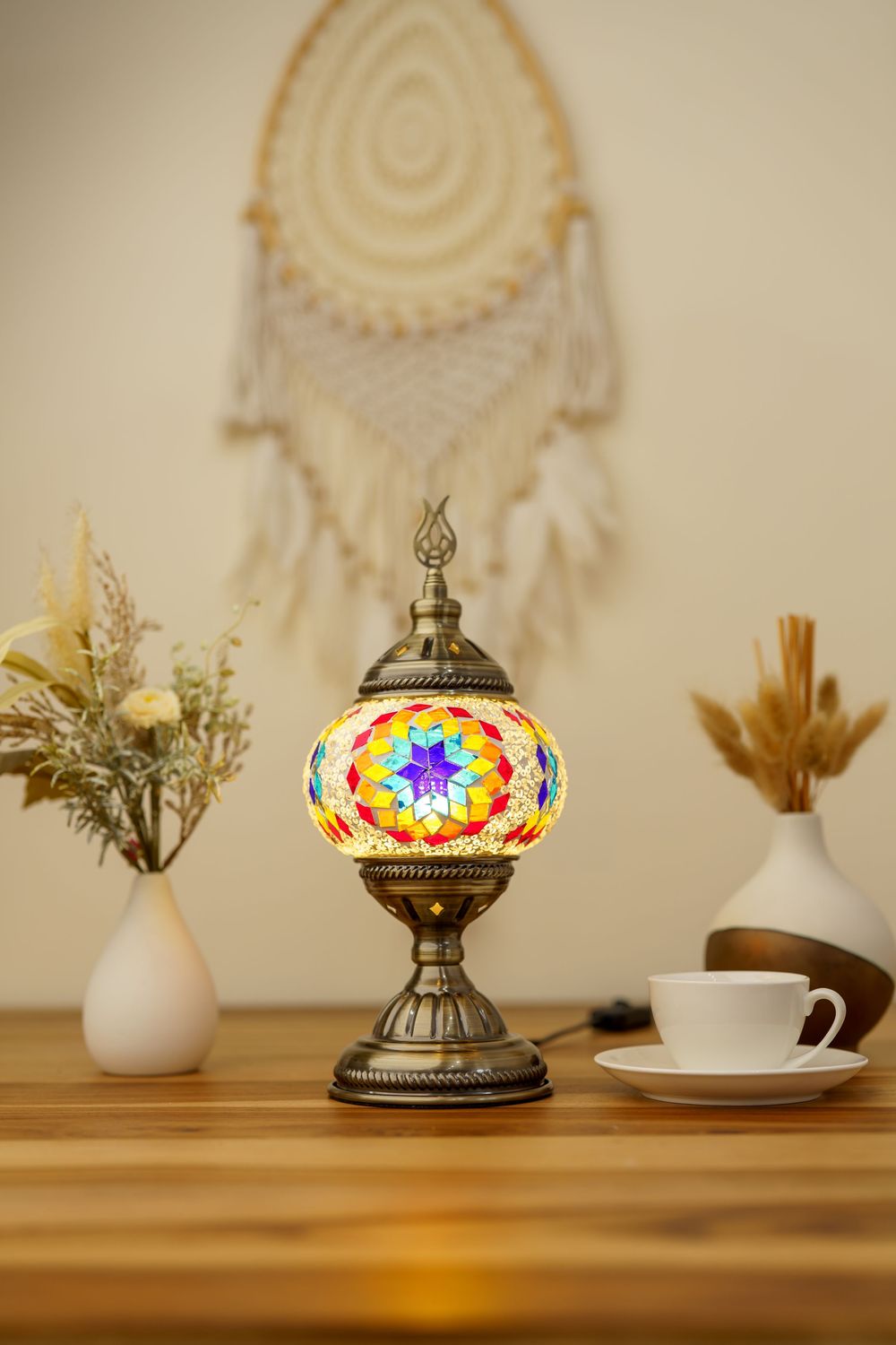 A colorful mosaic Turkish table lamp on wooden surface with dreamcatcher and coffee cup in background.