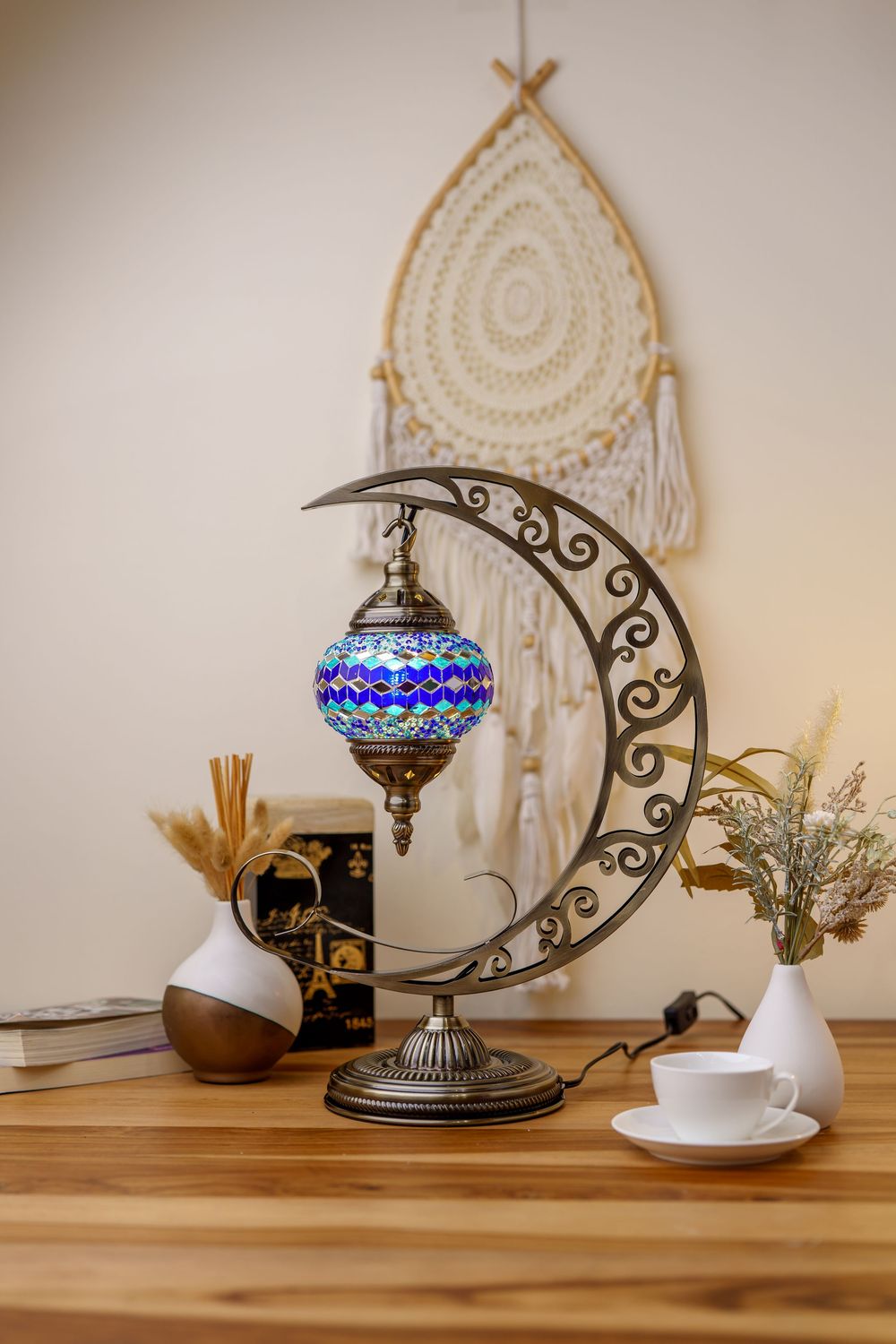 Turkish crescent moon lamp with blue mosaic glass shade displayed with dreamcatcher and white vases on wooden table.