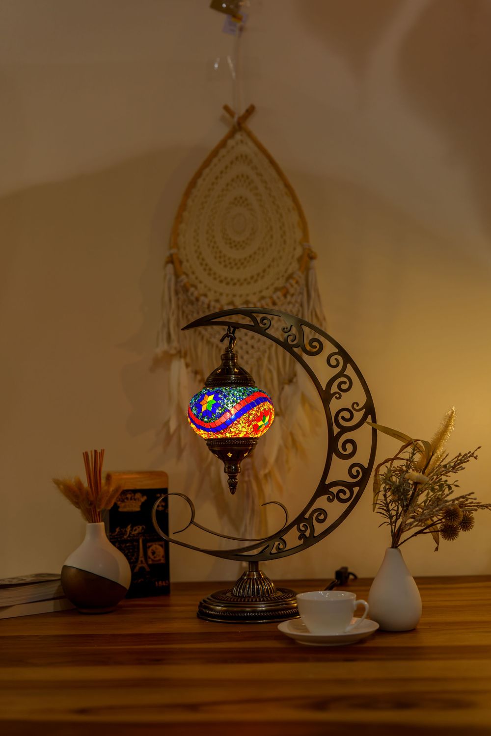 Illuminated mosaic crescent moon lamp glows against macrame wall hanging in dark room setting.