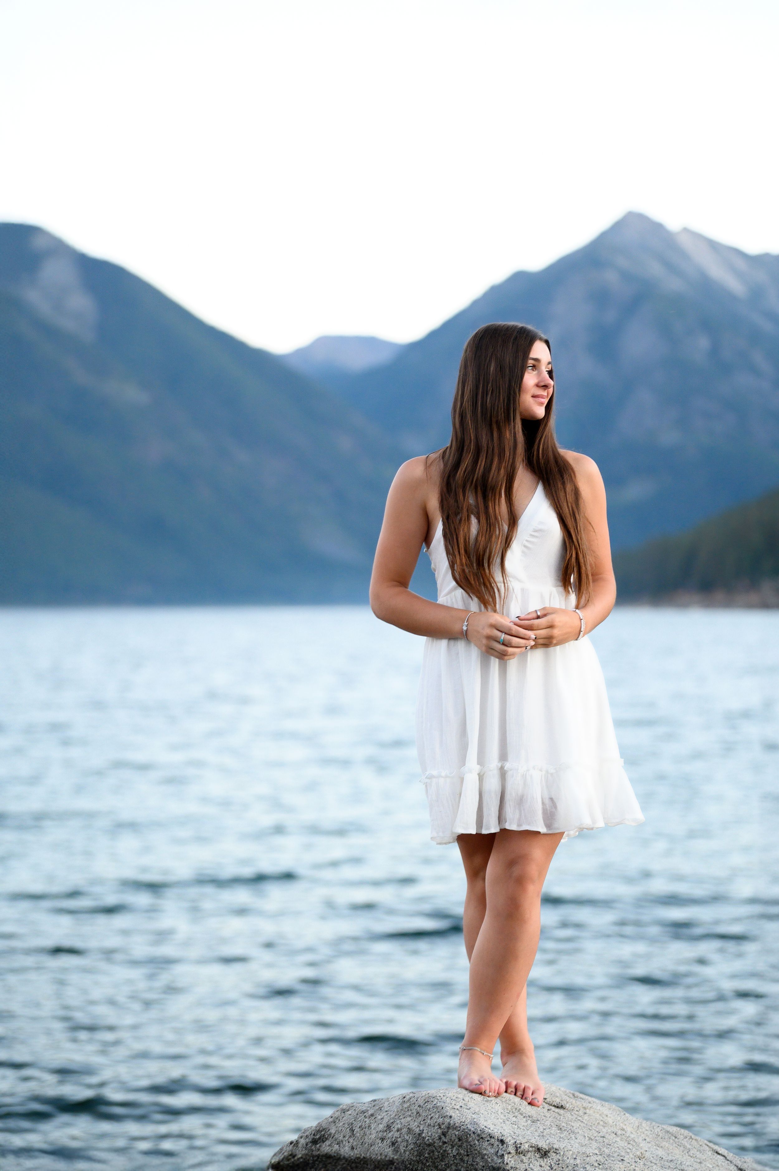Person in white summer dress stands barefoot on boulder overlooking mountain lake vista. Wallowa Lake, Joseph, Oregon