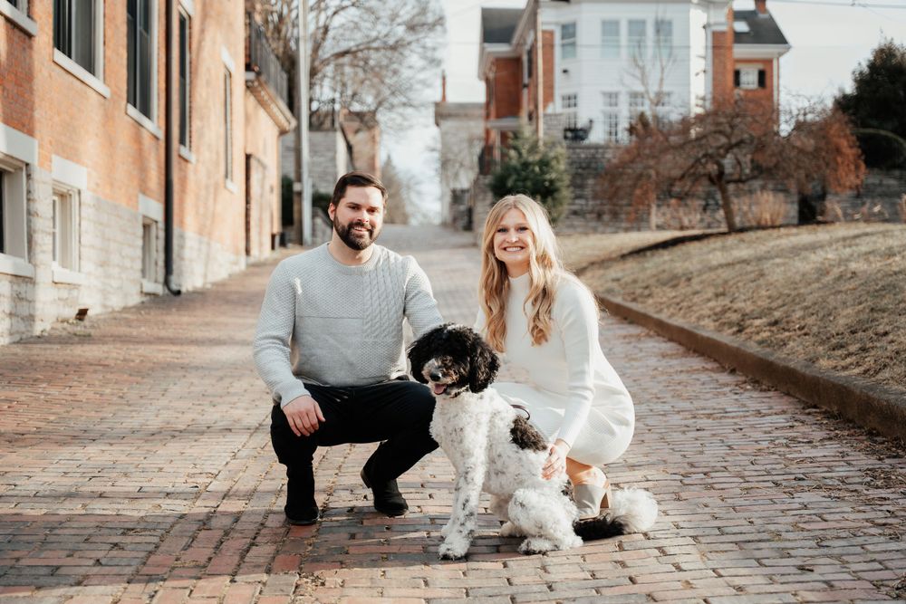 A couple crouches on a brick street with their black and white dog, set against a backdrop of historic buildings.