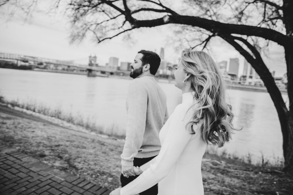 A couple in black and white stands by a riverside path beneath a tree branch, with the woman's hair flowing in the wind.