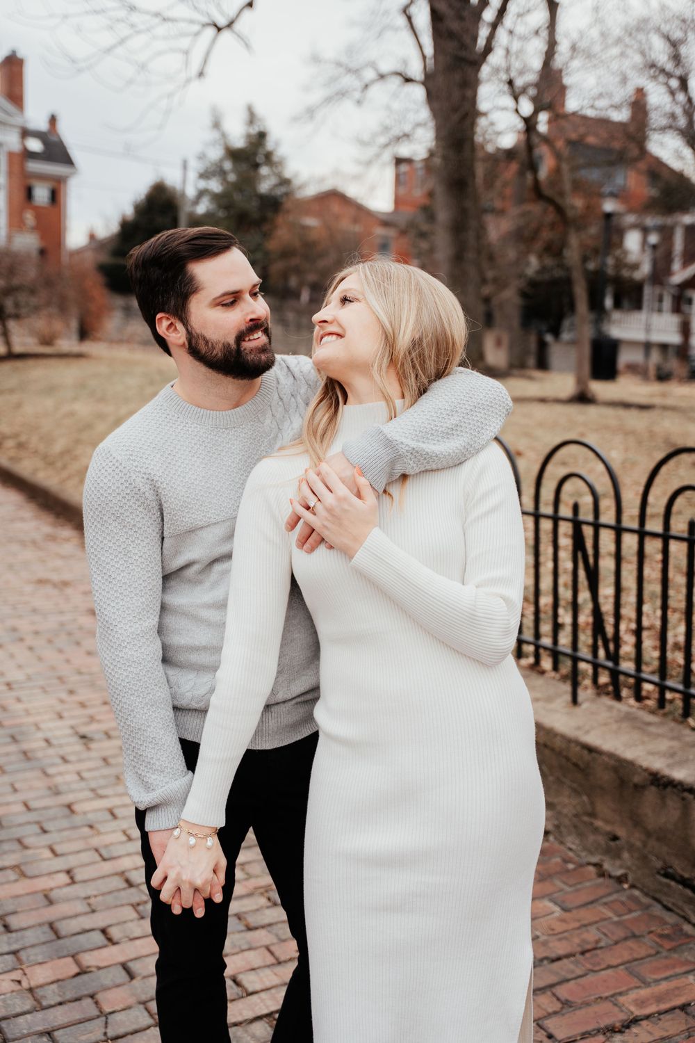 A couple in casual winter clothing embrace on a brick pathway outside historic buildings on an overcast day.
