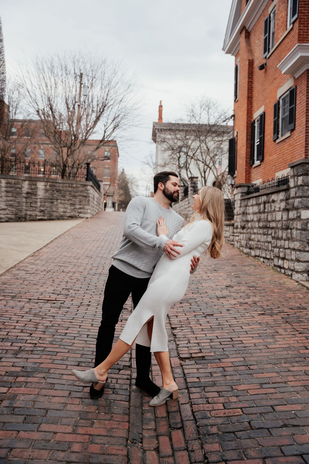 A couple dances romantically together on a brick sidewalk beside stone walls and historic buildings in an urban setting.